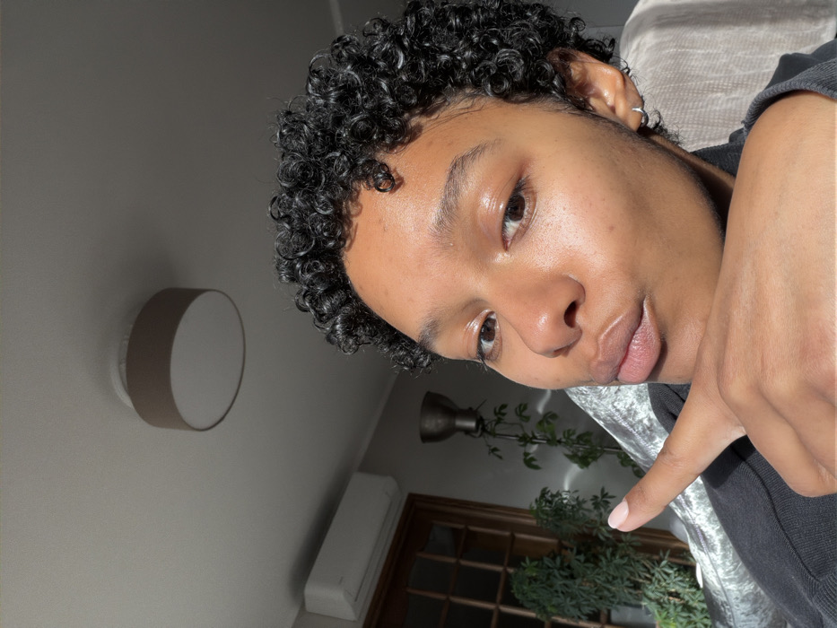 An indoor, eye-level photograph depicts an adult individual, appearing female, with dark skin and short, curly black hair, occupying the majority of the frame. The individual looks directly at the camera with a slight smirk and pursed lips. Their right hand is raised, with the index finger extended horizontally to the left and the thumb resting beneath the chin. A light-colored fingernail polish is visible on the index finger. A small silver hoop earring is present in the left ear. The subject wears a dark-colored garment, possibly a hoodie, partially visible on the right. Strong natural sunlight illuminates the right side of the subject's face and hair.
The background, an interior space, includes a light-colored ceiling with a round, drum-style ceiling light featuring a light brown fabric shade. A white, wall-mounted air conditioning unit is visible on the upper left wall, below which a wooden door with glass panels is partially visible. Further left, a tall, dark-colored floor lamp with a metallic, bowl-shaped shade stands near several green leafy plants. A textured, light-colored fabric, consistent with upholstery, is visible behind the subject's shoulder. The specific location within Champs-sur-Marne, France, cannot be determined from the visual evidence.