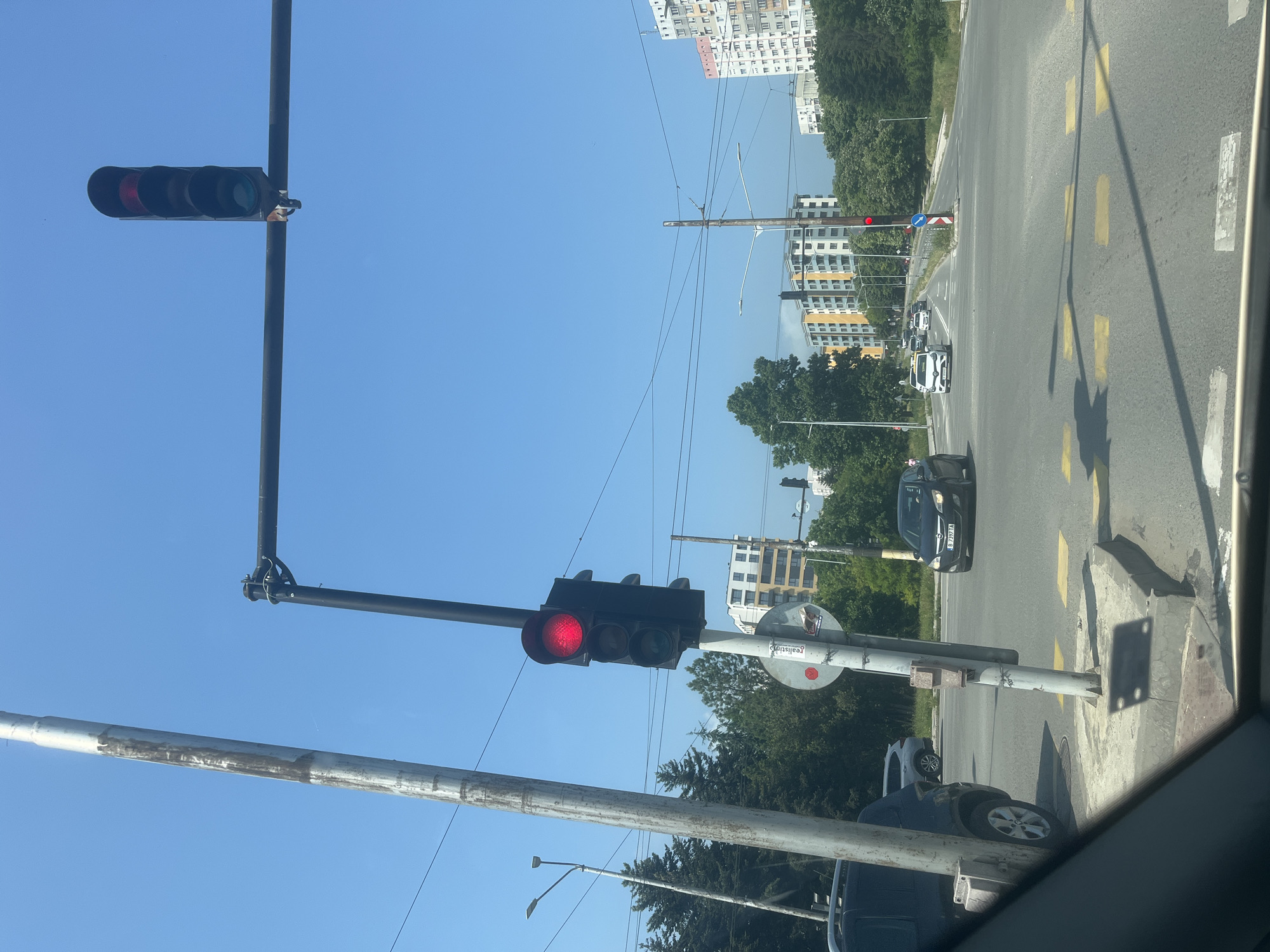 The image captures an urban intersection on a bright, clear day, viewed from inside a vehicle. The scene is oriented horizontally, showing the road stretching forward and to the right.
**Subjects:** The primary subjects are two traffic lights, both displaying red, indicating a stop for traffic. One traffic light is overhead, spanning across the road, while another is pole-mounted on the right side of the intersection. Several vehicles are visible, stopped at the red light. These include a black car in the foreground with a visible Bulgarian license plate "B 2377 KA", a white sedan further down the road, and other cars in the distance. The road itself is asphalt, featuring yellow dashed lane markings and white rectangular markings that likely indicate a crosswalk or stop line. In the background, multi-story residential buildings are interspersed with lush green trees. Utility poles, streetlights, and overhead wires are also prominent, set against a vast, clear blue sky. On a pole near the right-hand traffic light, a circular blue sign with a white arrow pointing right and downwards is visible.
**Activity or event taking place:** The scene depicts vehicles stopped at a red traffic light at an urban intersection, typical of daily city traffic.
**Notable details:** The time of day is clearly daytime, with bright sunlight and a cloudless blue sky, indicating fair weather. The architecture of the buildings and the license plate hint at an Eastern European location, consistent with Varna, Bulgaria (the provided context).
**Visible text:** The license plate "B 2377 KA" is visible on the black car in the foreground. On a pole-mounted sign, partially obscured text reads "realistig" or "realistiq".