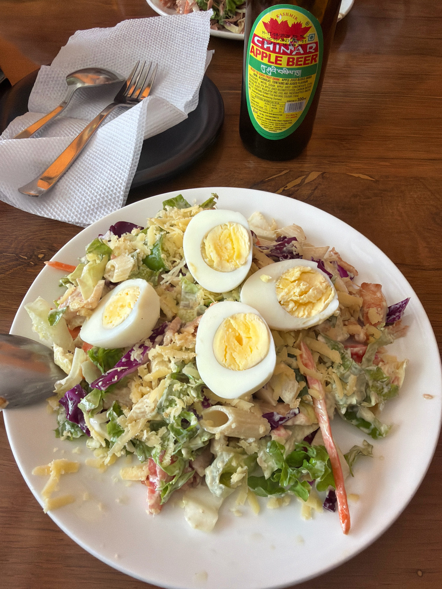 A close-up, top-down shot captures a white plate of salad on a wooden table. The salad features lettuce, shredded cheese, chopped tomatoes, red cabbage, pasta, and three halves of hard-boiled eggs. A fork and spoon rest on a folded white napkin to the left of the plate. To the right of the plate, a green glass bottle of "Chinar Apple Beer" stands upright. The bottle label is partially visible, showing text in English and what appears to be Tibetan script. The background is out of focus, but hints of another plate with food can be seen at the top. The overall scene suggests a meal being served in a casual dining setting. The presence of "Chinar Apple Beer" with Tibetan script on the label, combined with the overall aesthetic, is consistent with the city of Leh, India, where such local beverages are commonly found and served in restaurants and cafes.