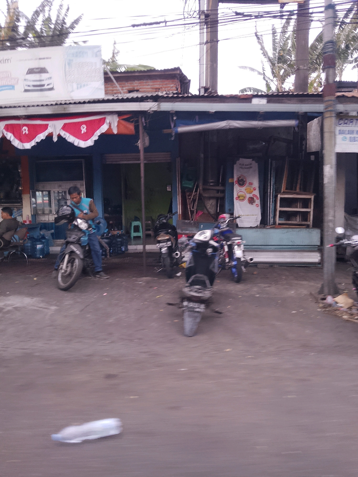 The image depicts a street scene in Malang, Indonesia, in front of a row of small shops. A man in a blue vest and jeans is sitting on a motorcycle, holding a helmet. Several other motorcycles are parked nearby, one of which is being operated by another individual further down the street. Large blue water bottles are stacked in front of one of the shops. Banners with text and images are displayed on the buildings. The ground is paved with asphalt, and a discarded plastic bottle is visible in the foreground. The overall atmosphere suggests a busy commercial area with transportation and retail activities.