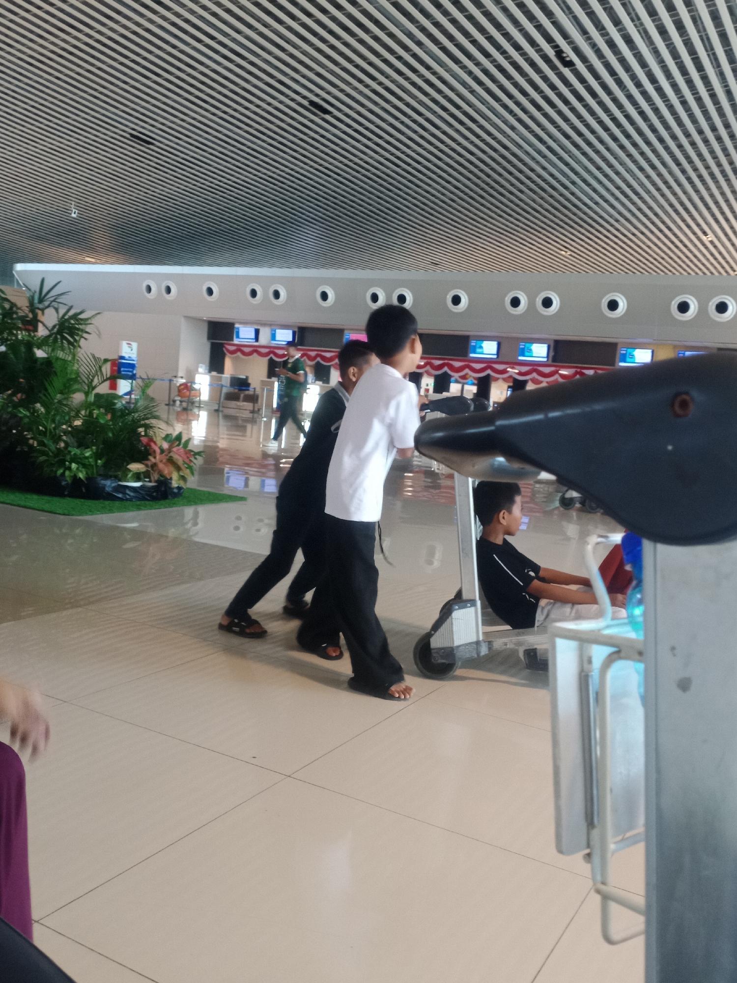 The image captures a scene within an indoor public space, likely an airport or a large terminal building, given the architecture of the ceiling and the presence of luggage carts. The floor is tiled with large, light-colored square tiles. In the foreground, a dark, bird-shaped object, possibly part of a decorative element or a cart attachment, is prominent on the right side.
Several individuals are present. In the middle ground, two boys, one in a white t-shirt and black pants, and another in a black t-shirt and black pants, are interacting with a luggage cart. The boy in white appears to be pushing the cart from behind, while the boy in black is partially obscured by the cart. To the left of them, another individual, possibly an adult, is seen from the waist down, wearing a dark garment. Further in the background, a young boy in a black t-shirt and shorts is seated on a luggage cart, looking to the side. Another person in a green shirt is walking in the far background.
The environment includes rows of overhead lights and circular decorative elements on a light-colored ceiling. On the left, a large arrangement of green plants, some with reddish leaves, is placed on a green mat, suggesting an attempt to bring nature indoors. Behind the plants, signage and information displays are visible, further supporting the idea of a public transportation hub. The overall impression is one of a busy transit area. The specific location in Baji Mangngai, Indonesia, is inferred from the prompt but not visually evident within the image itself.