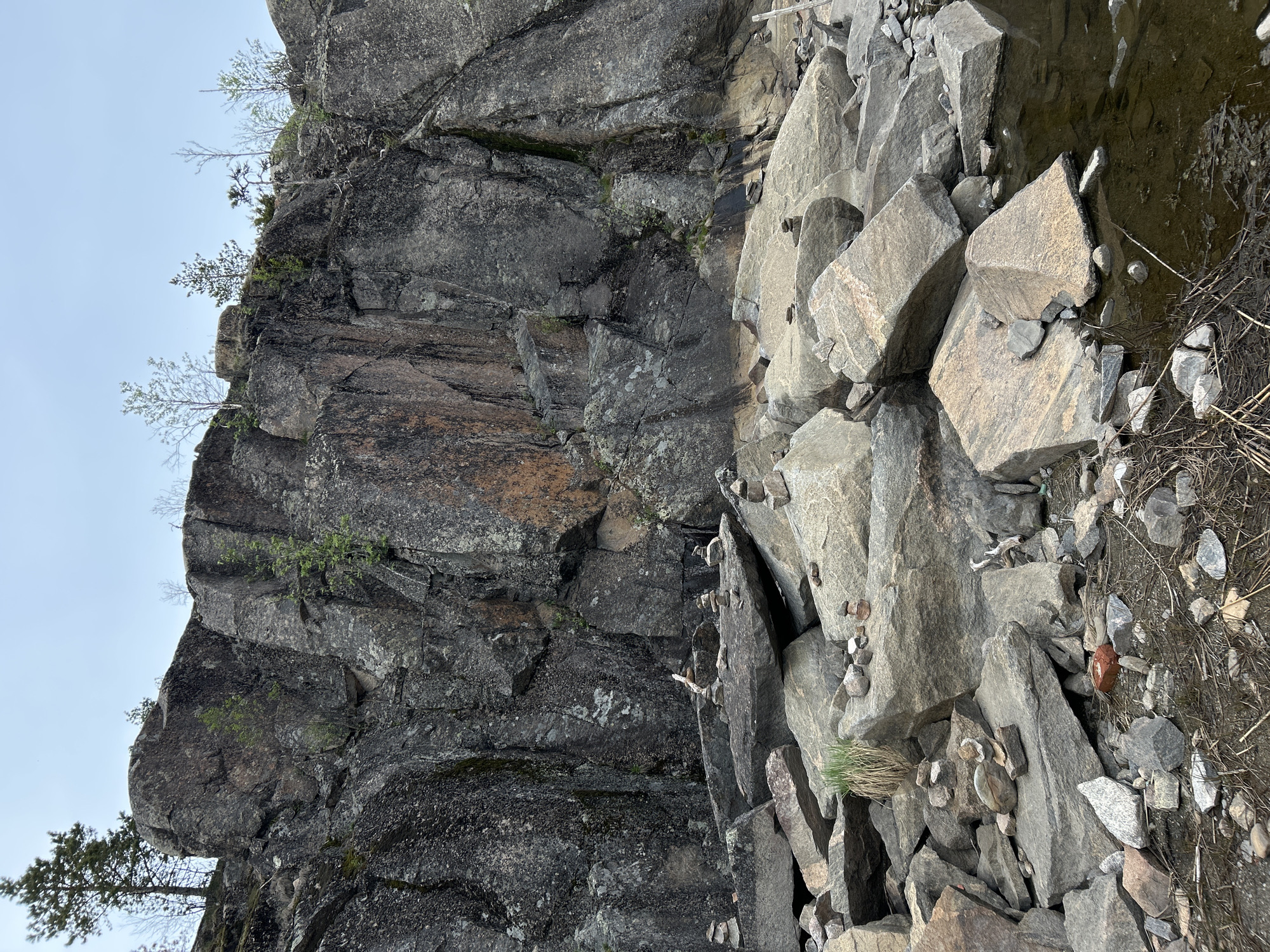 The image shows a rock formation. The rock formation consists of a cliff face with some small trees growing on it, and a pile of large, angular rocks at the base of the cliff. The foreground of the image contains smaller rocks and some sparse vegetation. The sky is visible in the upper left corner, and is a light gray-blue color. The scene appears to be in a natural environment. Location in the city of Lamberg, Russia is not apparent.