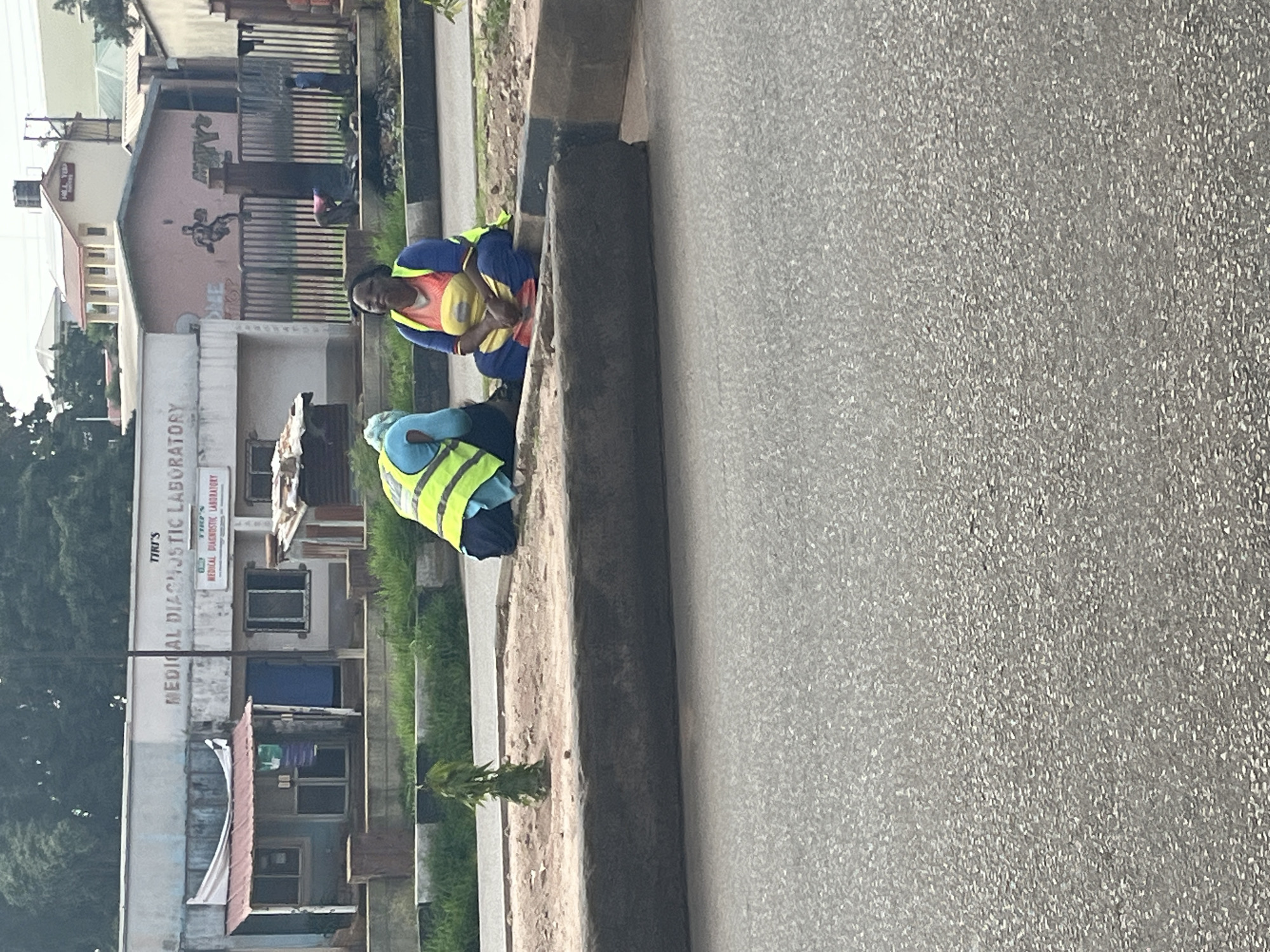 In Gura Top, Nigeria, two individuals, possibly sanitation workers due to their high-visibility vests, are seated on a curb next to a paved road. The person on the left wears a blue shirt and a yellow vest, while the person on the right wears a yellow top with blue trim and a yellow vest. Behind them is a low-lying building with signage that reads "TIRI'S MEDICAL DIAGNOSTIC LABORATORY." A small, leafy tree is planted in a patch of dirt to the left of the seated individuals. The environment suggests a commercial or service area within the city.