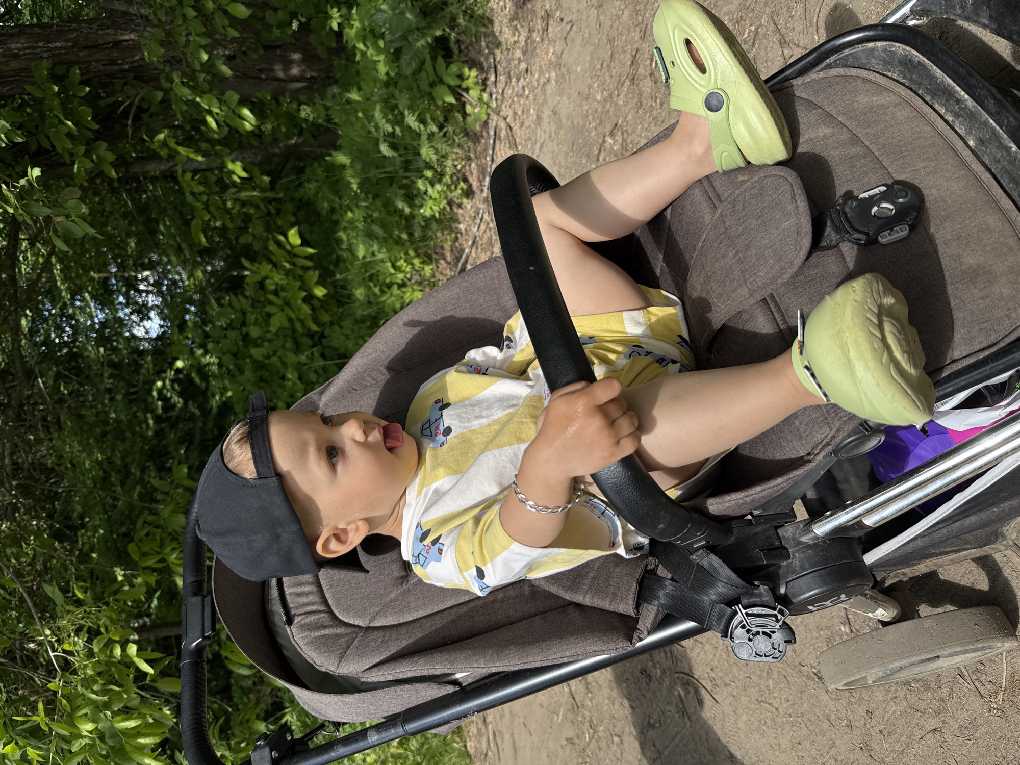 A young child is seated in a brown and grey stroller, outdoors on a sunny day in what appears to be Rusanovo, Russia. The child, likely a toddler, wears a black baseball cap reversed, a yellow and white striped t-shirt adorned with car motifs, yellow shorts, and light green clogs. A silver chain bracelet is visible on their right wrist. Both hands grip a black handlebar, which is part of the stroller's design, suggesting an interactive element. The child has an open mouth with their tongue slightly visible and looks towards the left of the frame with an expression of curiosity or engagement.
The stroller features a safety harness and visible parts of its frame and wheels resting on a dirt path. Surrounding the stroller and filling the background is abundant green foliage, including trees and bushes, indicating a natural, perhaps wooded, outdoor environment. Bright daylight illuminates the scene, creating clear contrasts and highlighting the details of the child and their surroundings. The overall impression is a cheerful outdoor outing. No legible text is visible within the image.