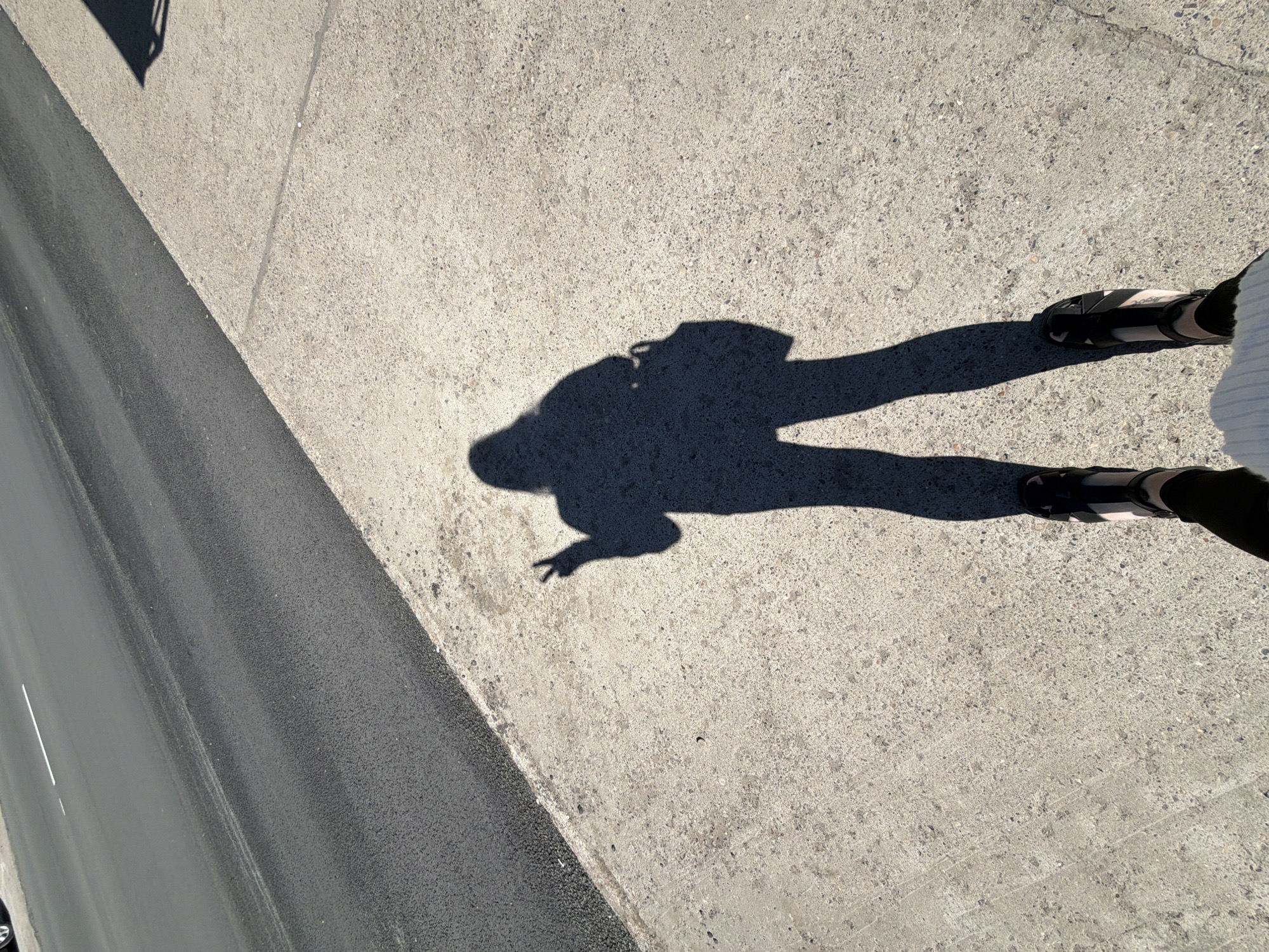 The image depicts a shadow of a person standing on a sidewalk next to a road. The shadow suggests the person is wearing sandals and possibly holding up two fingers. The road is asphalt. The sidewalk appears to be made of concrete. The overall scene appears to be outdoors in daylight. There is no indication that the image was taken in Chaikovsky, Russia.