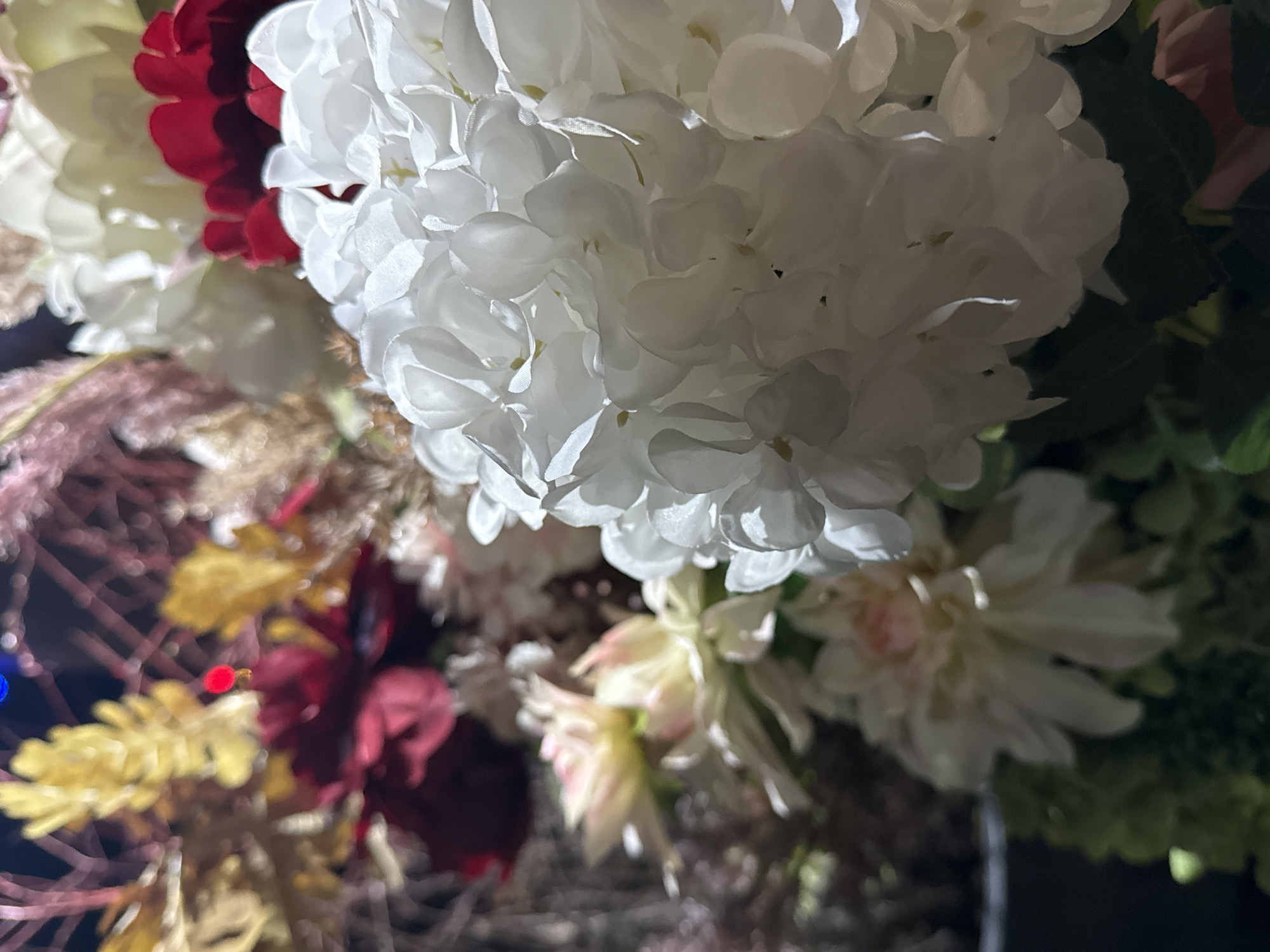 The image shows a close-up view of an arrangement of artificial flowers. Predominantly, there are large white flowers, likely hydrangeas, dominating the frame. Red and cream-colored flowers are visible as well, adding visual interest and texture. The foreground features gold or tan foliage. The background includes green foliage. The scene is dimly lit. The location is not discernible from the image itself.