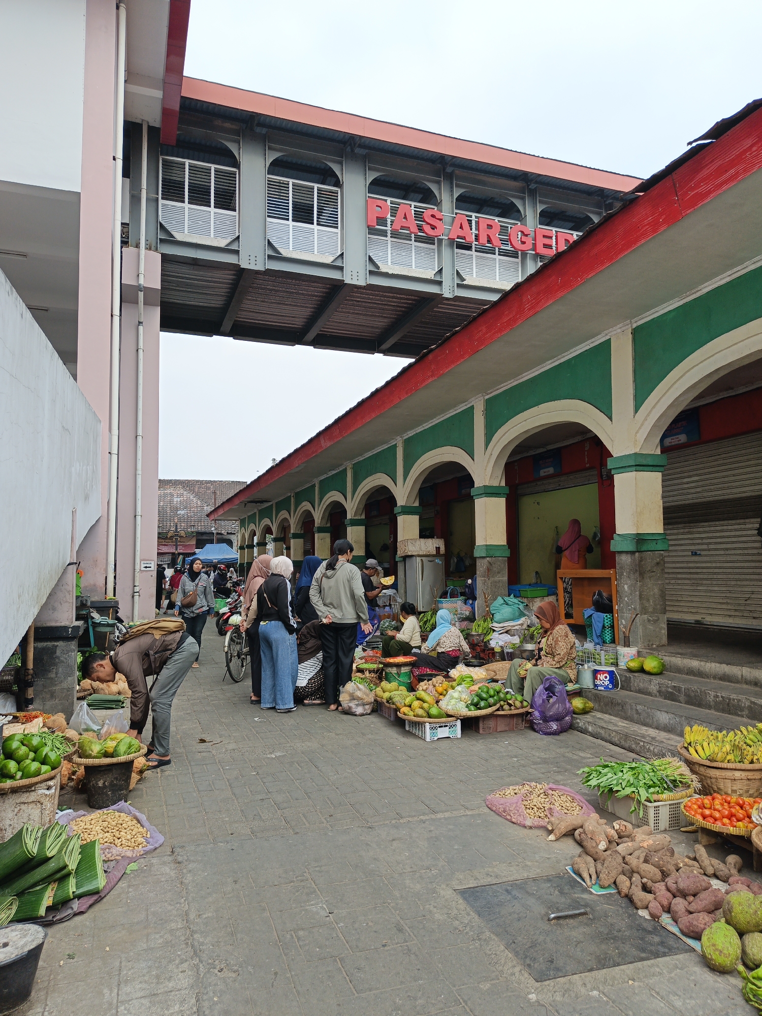 The image shows the outdoor market area in front of the Pasar Gede building in Klaten, Indonesia. The Pasar Gede building has a distinctive red roof and green archways, with a pedestrian bridge overhead bearing the red lettering "PASAR GEDE". Several market stalls are set up on the paved ground, displaying a variety of fresh produce including bananas, tomatoes, sweet potatoes, avocados, peanuts, and what appear to be bundled green vegetables. People are seen browsing and shopping at the stalls. Some individuals are wearing traditional Islamic head coverings. A bicycle and a motorcycle are also visible, suggesting transportation within the market area. The overall scene depicts a bustling local marketplace.