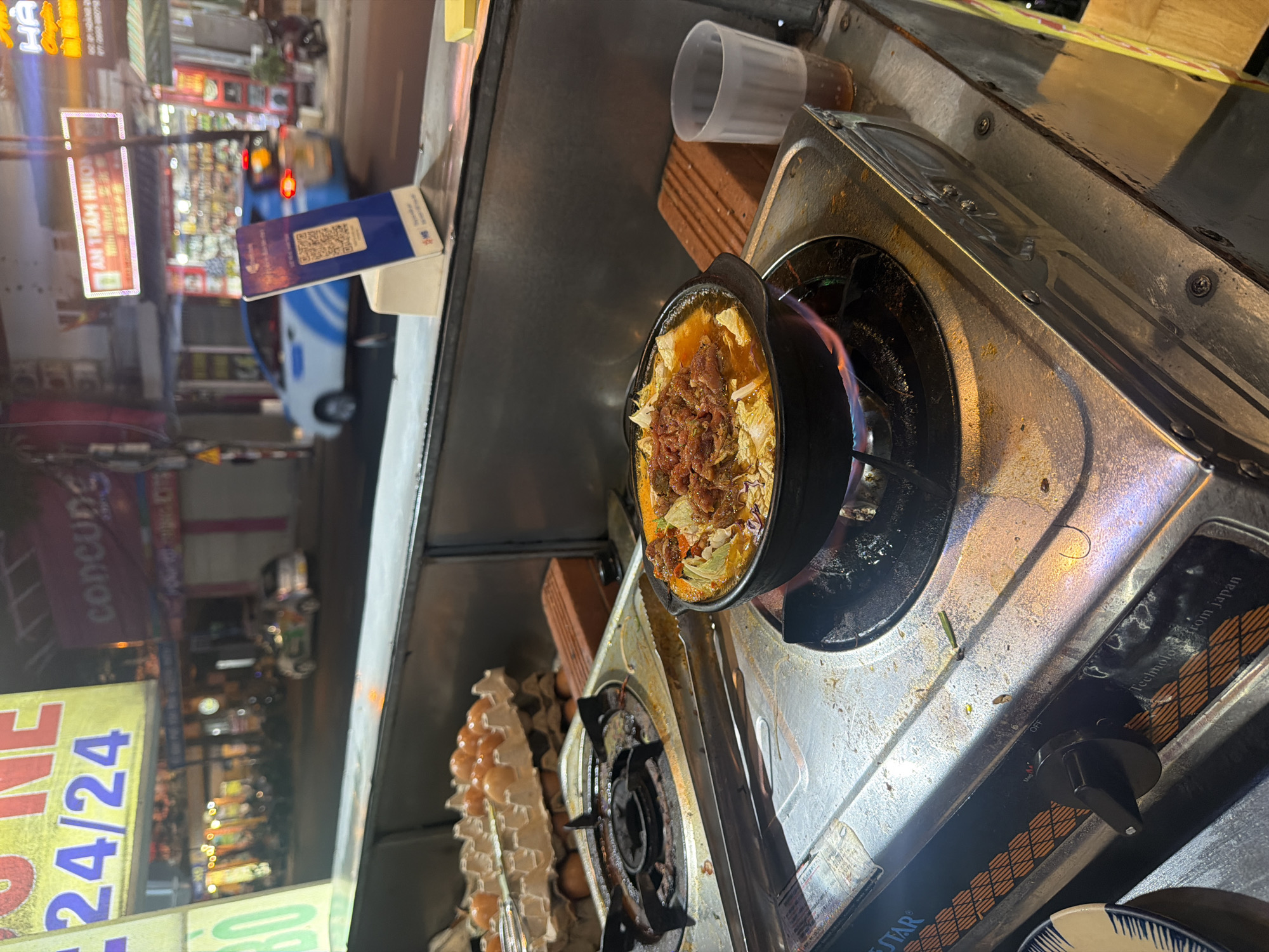 This image depicts a nighttime street food scene within an urban area of Nha Trang Ward, Vietnam.
In the foreground, a stainless steel food stall counter hosts two portable gas stoves. The stove on the right is actively heating a black ceramic pot containing a simmering dish, identifiable by leafy greens and reddish-brown meat (possibly beef) on top, over a visible blue and orange flame. Adjacent to the pot on the right, a clear ribbed plastic cup holds a dark liquid beverage with ice. Between the two stoves, a pair of metal tongs rests on the counter. To the left of the stoves, multiple cardboard cartons stacked vertically contain numerous brown eggs. A blue and white stand displaying a QR code for mobile payment, featuring the "MB" logo, is prominently positioned on the counter near the center-right. A reddish-brown brick is placed to the right of the active stove. The counter surface shows signs of use with some residue. The second stove on the left is inactive but appears ready for use. Text visible on the inactive stove reads "BLUE STAR" and "Technology from Japan."
The background, largely out of focus, reveals a bustling street scene under artificial illumination. Commercial buildings line the street, adorned with various neon and backlit signs displaying Vietnamese text. Notable signs include a prominent yellow sign on the far left with "24/24" (indicating 24-hour service), a pink sign further back reading "concung," and a rectangular white and red sign in the mid-ground stating "AN TRẬM HƯƠNG." Vehicles are present on the street; a white car with blue stripes, characteristic of a ride-sharing service, moves from right to left in the mid-ground. Another smaller white vehicle with green branding is visible further down the street on the left. Motorbikes are also discernable. No individuals are clearly identifiable in the blurred background.