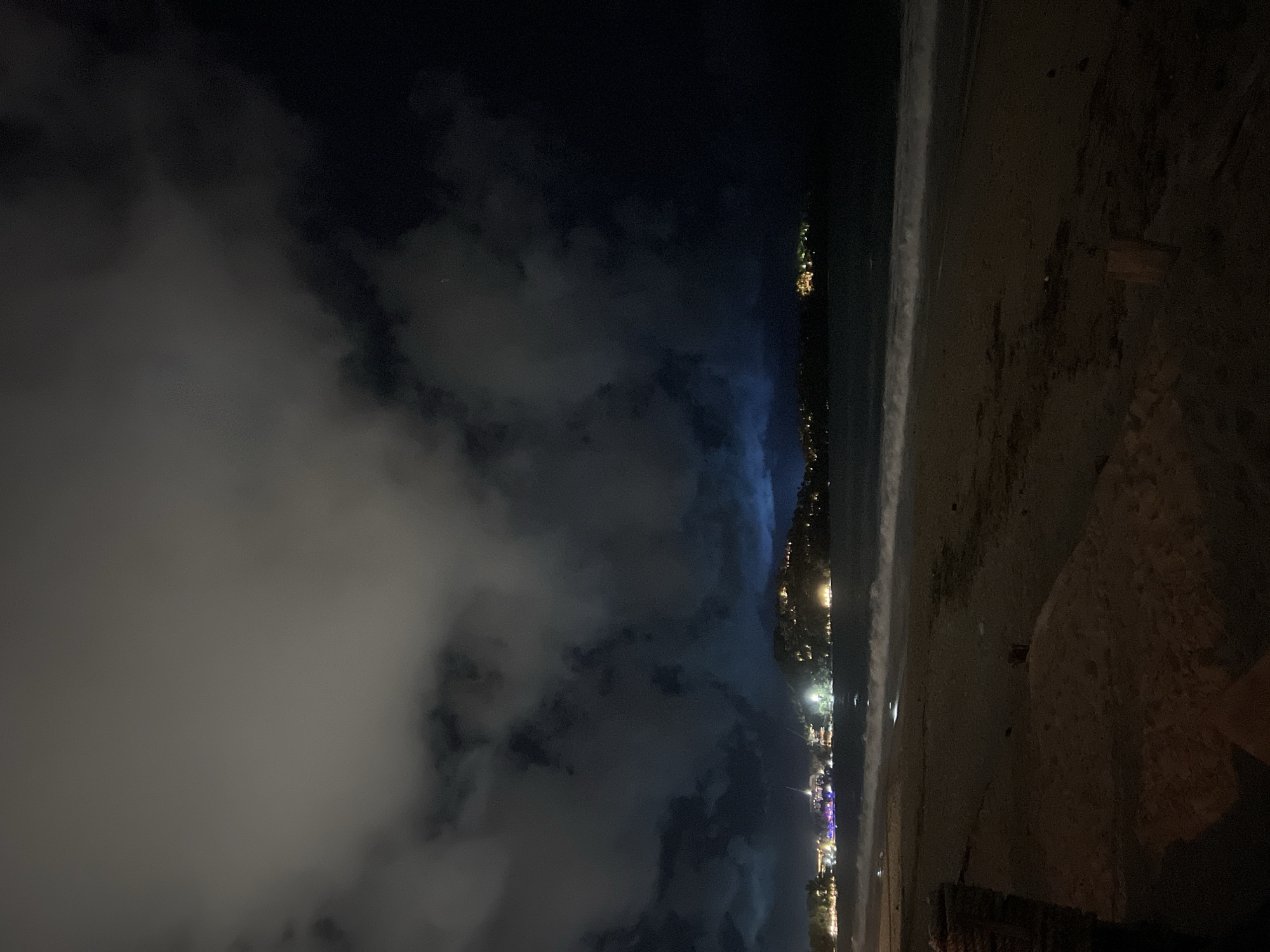 The image depicts a beach at night. The scene is dimly lit, with city lights visible along the coastline. The sand is dark and extends into the foreground. Clouds fill the sky. The city is unknown.