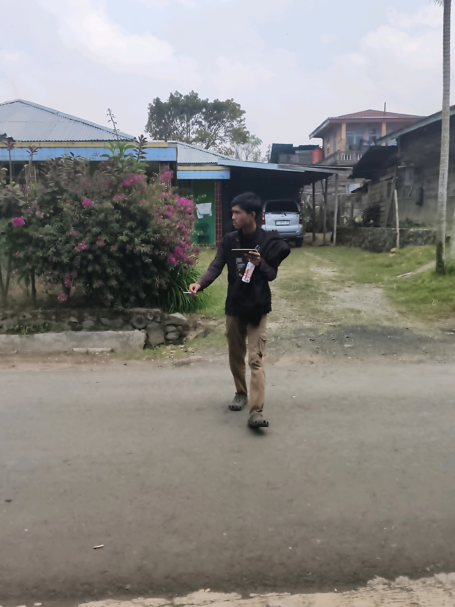 A young man with dark hair is walking on a paved road in front of some residential buildings and a dense bush with pink flowers. He is wearing a black long-sleeved shirt, khaki cargo pants, and dark clogs. In his right hand, he holds a smartphone and a white bottle with red lettering. His left arm is bent at the elbow, and his hand is reaching towards his side. Behind him, a silver SUV is parked under a carport attached to a building. Further in the background, there are more houses and trees. The sky is overcast. This scene appears to be located in a residential area within Lindung Jaya, Indonesia, characterized by modest housing and lush vegetation.
