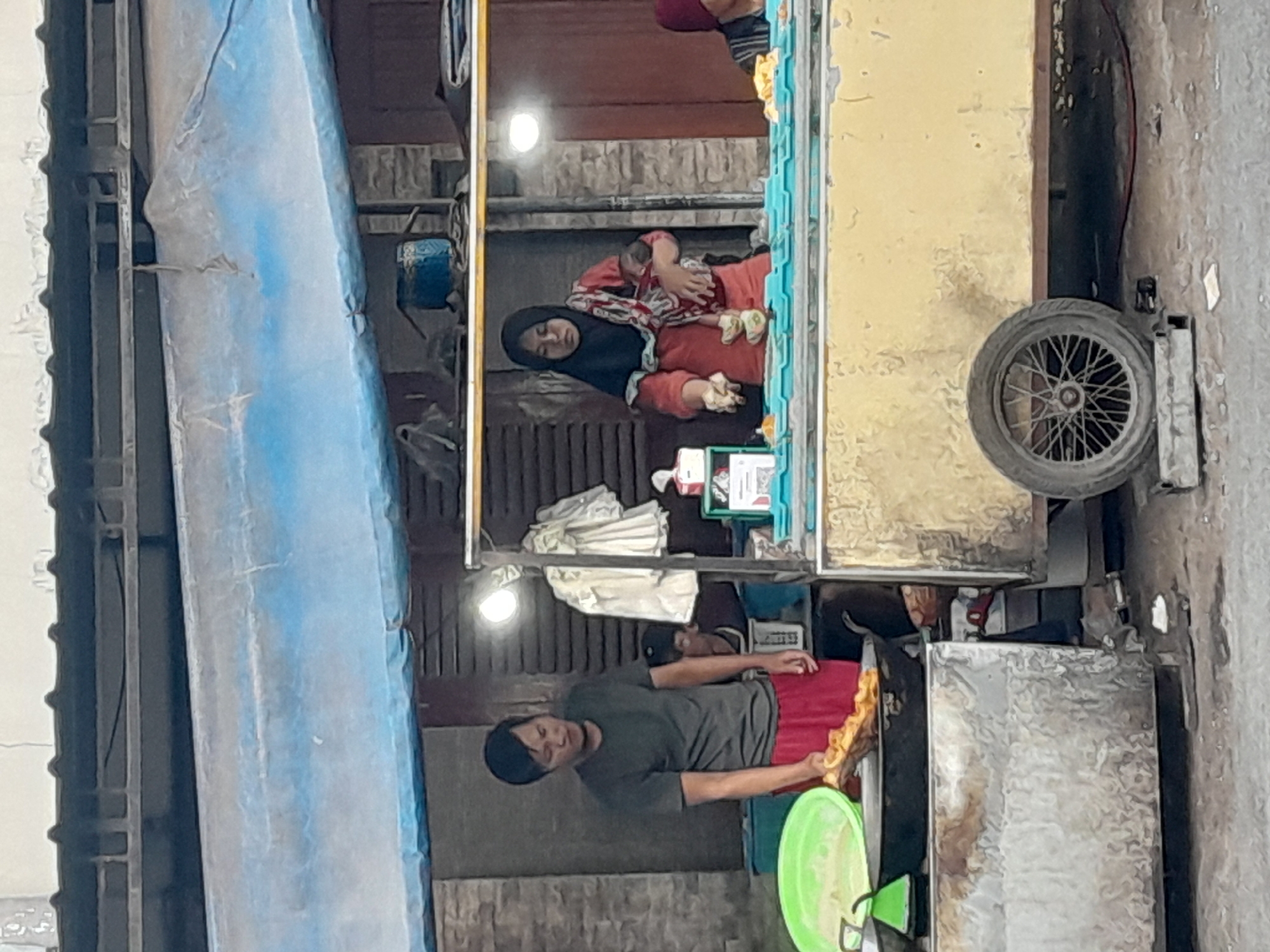 This image depicts an outdoor food stall in Gentengwetan, Indonesia, likely a street-side vendor or market. The stall appears to be a portable cart with a yellow exterior, fitted with a wheel for mobility. Above the stall, a blue tarp is stretched across, providing shade.
In the foreground, a man in a dark t-shirt and red shorts is actively engaged in preparing food. He holds a long, golden-brown fried item over a large black wok, which is situated within a large green bowl. To the right of the man, a woman wearing a hijab and an orange-red outfit is seen holding a baby. She is likely also involved in the food preparation or sales. Another person, possibly an employee, is partially visible behind them.
The environment suggests a bustling area, with hints of a building structure behind the stall and a textured ground surface, indicating a paved or dirt area typical of a street or market. The lighting suggests it may be daytime, with artificial lights also visible on the stall. The overall scene captures a snapshot of daily commerce and life in the Indonesian town.
