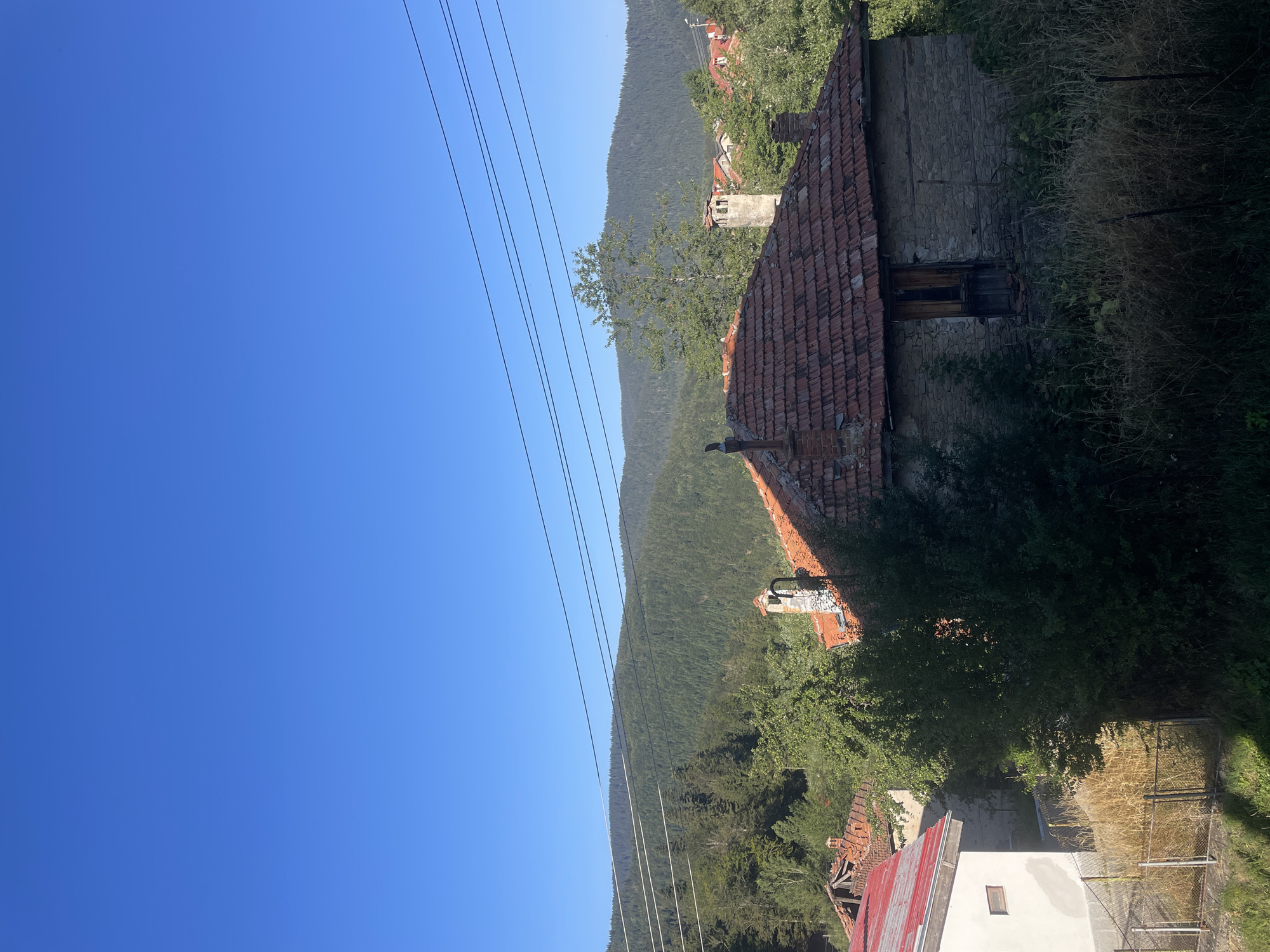 A rural mountainous landscape with houses in Manastir, Bulgaria, is depicted under a clear blue sky. The foreground features a weathered stone house with a red tile roof and a dilapidated wooden door. To the left, a white house with a red metal roof is visible. Behind these structures, dense green forests cover rolling hills, with power lines strung across the scene. The overall impression is one of a serene, somewhat rustic, mountain village.