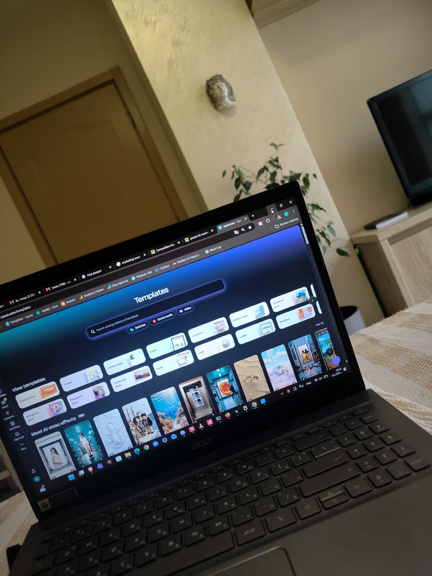 An indoor, eye-level shot captures an open Acer laptop resting on a textured, light-colored surface, possibly a blanket or bedspread. The laptop's screen displays the Canva website, specifically the "Templates" section. The main heading "Templates" is prominent, with a search bar below it reading "Search among millions of templates." Filter buttons for "Business," "Social media," and "Video" are visible.
A sidebar on the left shows categories like "View templates," "Presentation," "Poster," "Instagram post," and "Instagram Story," along with "Meet AI video effects New." Various template thumbnails are displayed, including examples for "Email," "Invitation," "Code," "Logo," "Leaflet," and "Brochure." The browser tabs at the top of the screen show multiple open pages, including "М Вх. поща (2292)," "M Inbox (706)," "marketing analy," "Competitive Ma," "power-bi-marke," "Шаблони - Can," "canva.com/templates," "Templates - Canva," "Netlify - host," "domain," "Analytics | Home," "Общ преглед," "Telegram Ads," "Сървър," and "Workers & Pages." The laptop's taskbar shows "ENG" (English), the time "15:54," and the date "4.6.2026 г." (June 4, 2026). A "Full HD 1080" sticker is visible on the bottom left bezel of the screen. The keyboard features a Cyrillic layout.
In the background, a light-colored wall with some texture or minor discoloration is visible, along with a closed light brown door to the left. A small decorative bust or statue is mounted on the wall. To the right, part of a green leafy plant is seen, and further right, a dark television screen rests on a wooden cabinet. The overall setting suggests a casual home environment.