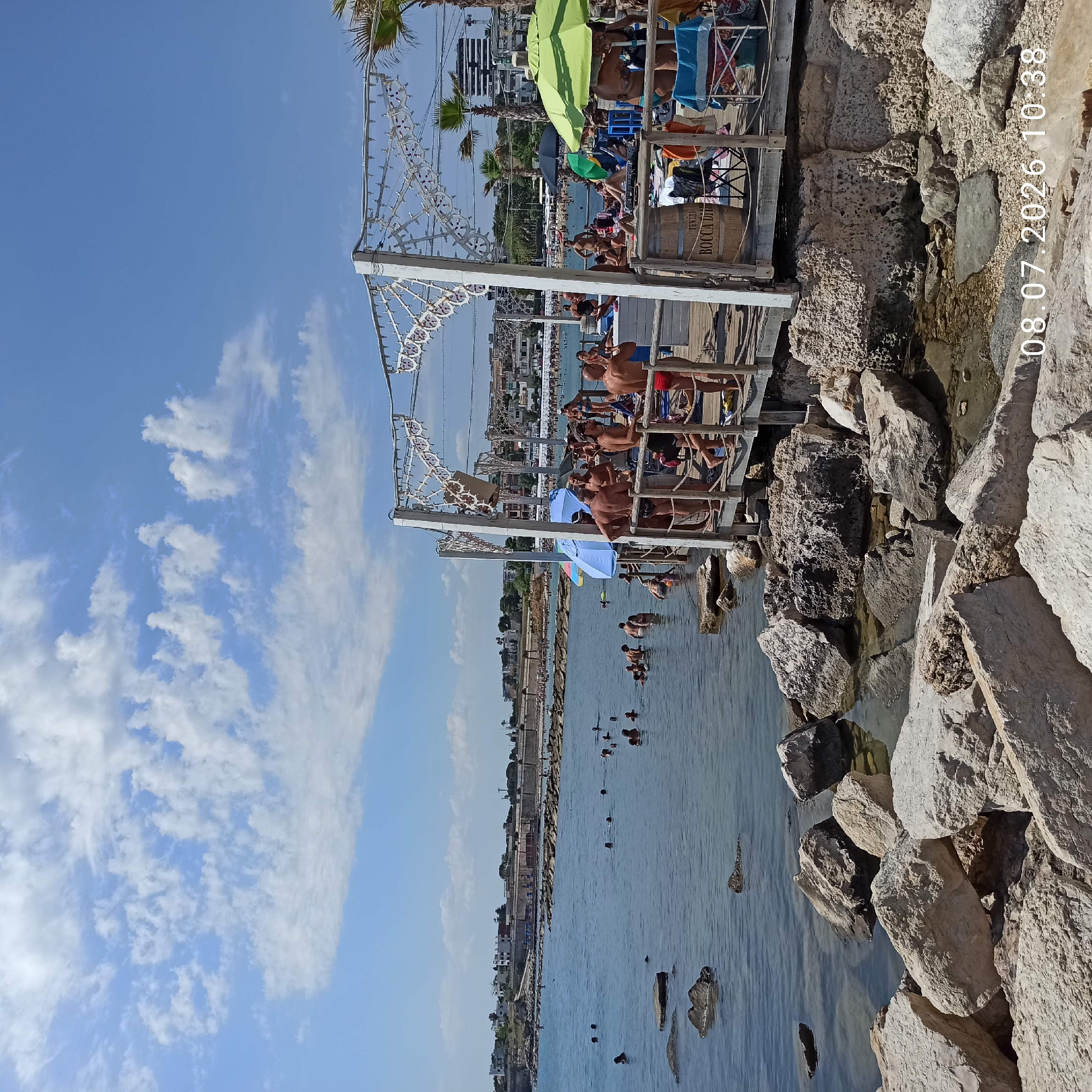 A daytime, eye-level, wide shot captures a lively scene at a beach in Trani, Italy. The foreground is dominated by large, rough, light-colored rocks, some submerged in clear, shallow water. Beyond these rocks, the sea stretches out, dotted with numerous people swimming and wading. A wooden pier juts out into the water, crowded with individuals socializing and relaxing. Some are standing, others are seated on chairs or loungers, and a few are near a bar area. Two large umbrellas, one blue and one green, provide shade. The background reveals a long stone wall lining the coastline, with buildings and palm trees visible beyond. The sky is a clear blue with scattered white clouds. The overall impression is one of a bustling summer day at a seaside resort. A date and time stamp, "08.07.2026 10:38," is visible in the lower right corner of the image.