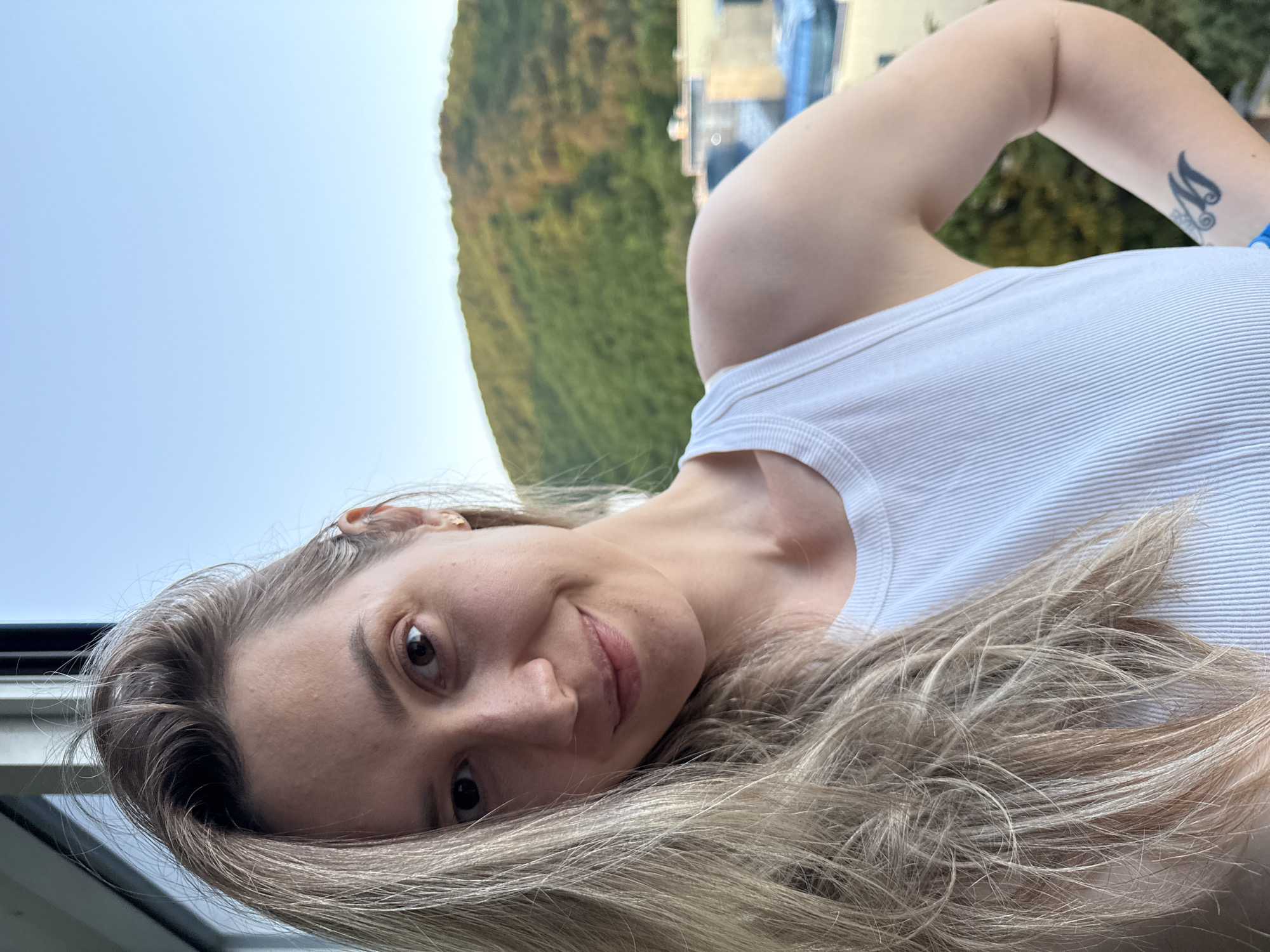 A young adult woman with long, ash blonde hair, wearing a white ribbed tank top, is captured in a selfie-style portrait in Nebug, Russia. She is positioned slightly off-center, gazing directly at the camera with a subtle smile. Her head is tilted slightly, and her right arm is bent with the elbow outward. A small, star-shaped earring is visible in her right earlobe, and a stylized "M" tattoo is present on her inner right forearm. The background features densely forested hills under a clear, light blue sky, with several low-rise buildings visible in the mid-ground below the hills. The lighting is soft and natural, suggesting late afternoon or early evening. A dark vertical window frame is partially visible on the far left edge of the image.