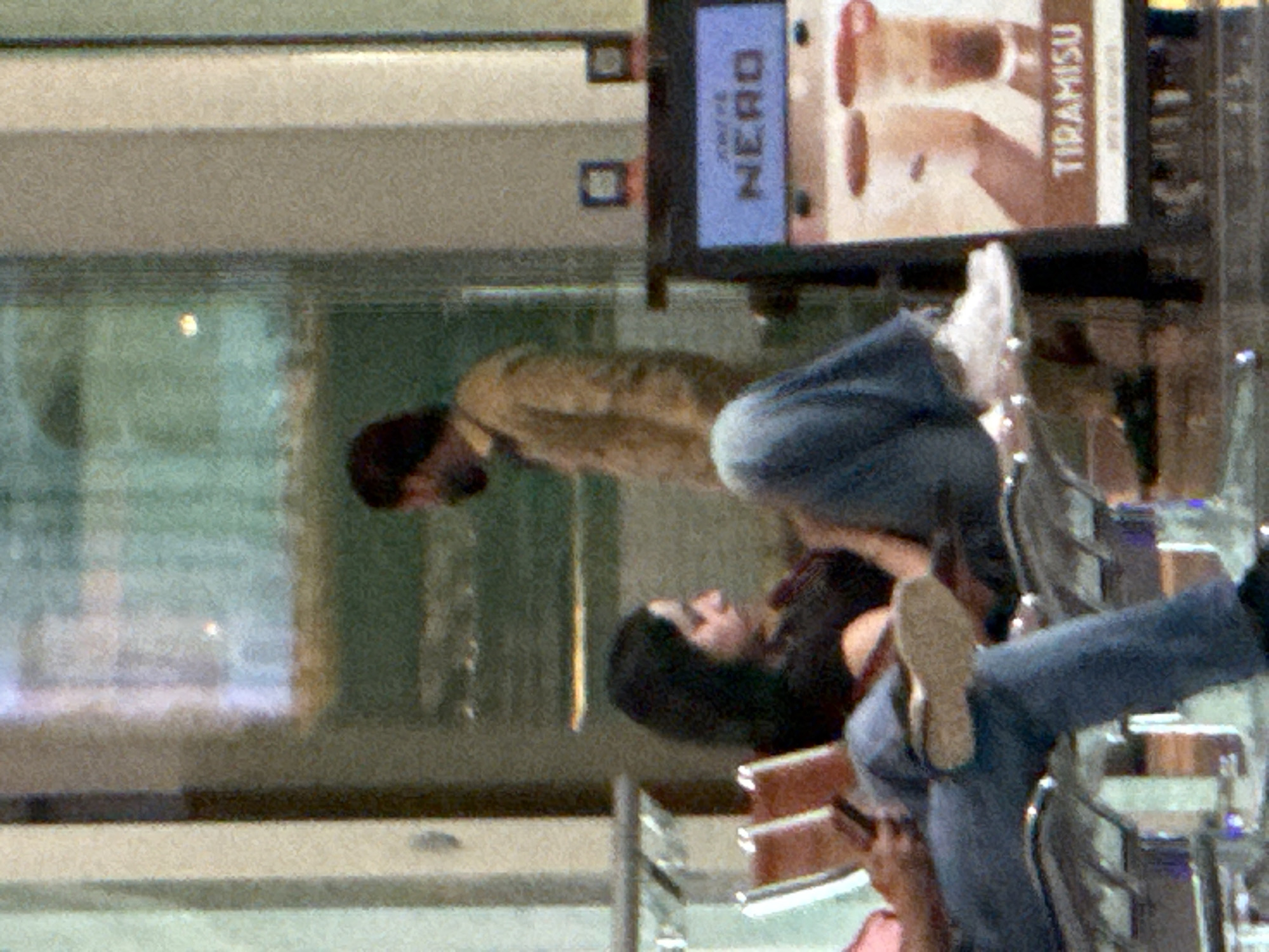 The image depicts an indoor scene, possibly an airport waiting area, in Seeb Province, Oman. In the foreground, a person wearing blue jeans and white sneakers is seated with their legs extended. To the left, two individuals are seated on chairs. One person, with dark hair and wearing a black top, is looking down, appearing to be resting. Further to the right, a man in a light brown jacket stands near a glass partition. In the background, a digital display screen shows the word "NERO" and an image of Tiramisu. The overall ambiance suggests a public waiting space with a food or beverage offering.