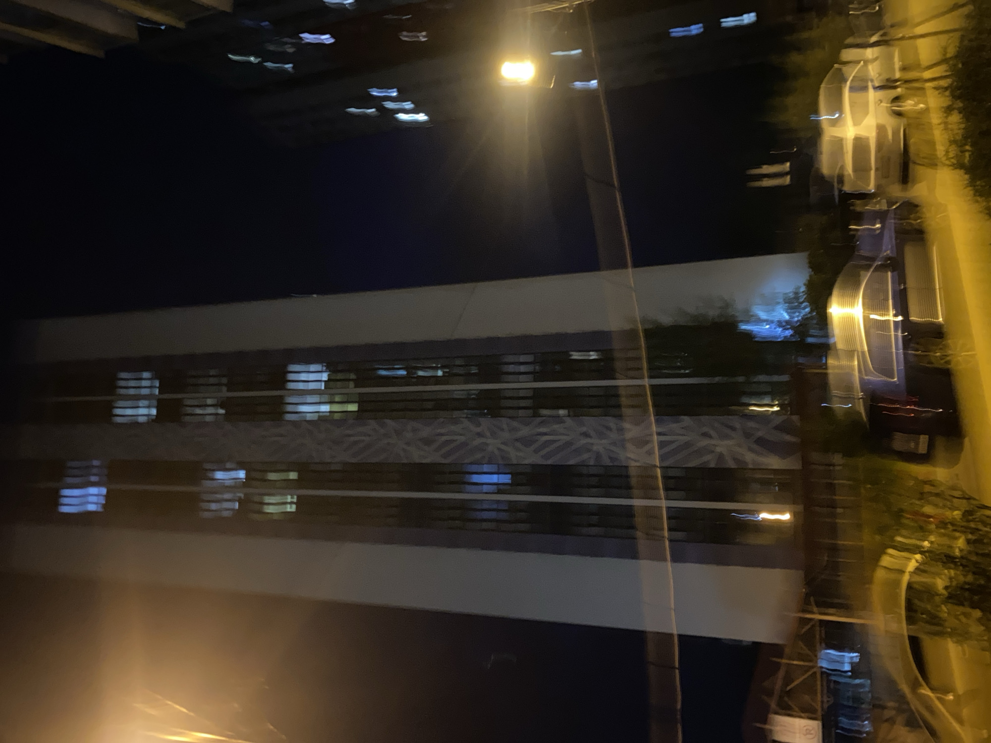 The image depicts a nighttime scene in Kyiv, Ukraine, likely in a city environment. The photograph is characterized by motion blur, suggesting it was taken from a moving vehicle or handheld with a slow shutter speed.
The primary subject is a long, modern building with numerous windows. Many of these windows are illuminated with a cool, bluish light, indicating interior lights are on. Some sections of the building's facade feature a patterned design that is partially visible through the blur. Above the building, in the dark night sky, several streaks of light are visible, possibly from distant traffic, lights on other buildings, or even fireworks. A prominent streetlamp with a bright yellow light is visible, casting a glow and creating light trails.
To the right of the building, there are blurred shapes that appear to be vehicles, possibly cars and a bus, suggested by their elongated forms and reflections of light. Trees or foliage are also present on the right side, their forms distorted by the motion and low light. The overall impression is one of urban activity at night, with the lights of the city creating a dynamic and somewhat abstract visual. There is no visible text in the image. The time of day is clearly night, and the weather is not discernible.