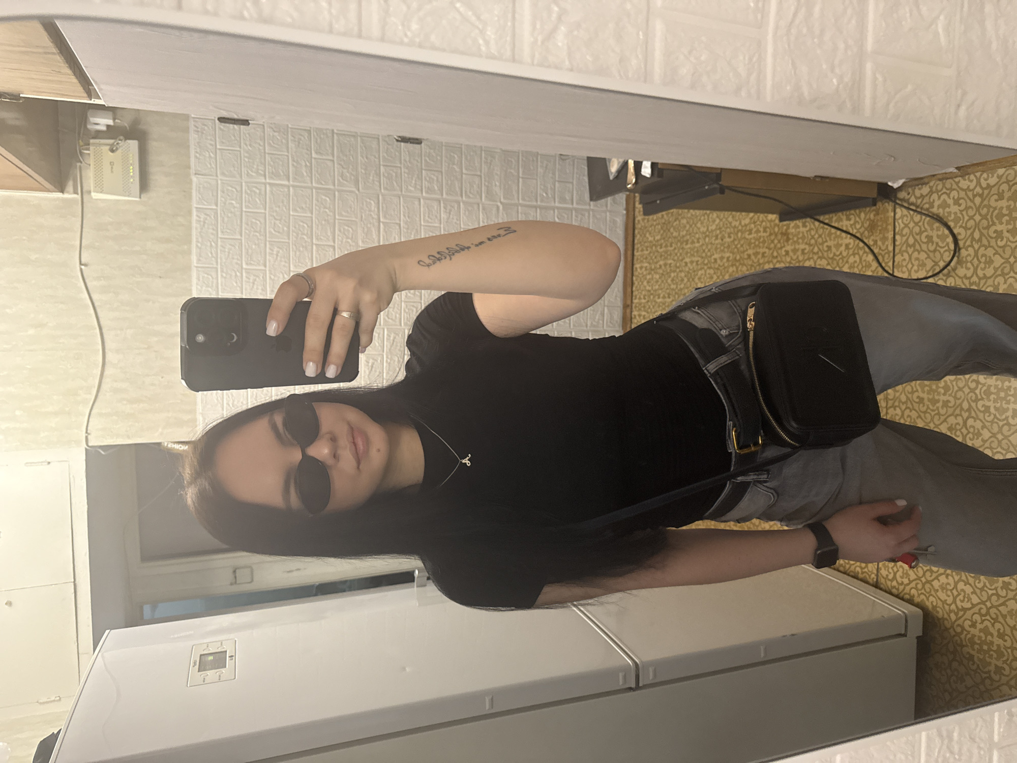 A woman is taking a selfie in a mirror. She is wearing sunglasses, a black t-shirt, gray pants, a black crossbody bag, and an Apple Watch. She has a tattoo on her left arm and is holding a black smartphone. The background includes a white brick-patterned wall, a yellow patterned floor, a cabinet, and a window. The location appears to be an indoor space, possibly in Mytishchi, Russia, but this cannot be definitively determined from the image.