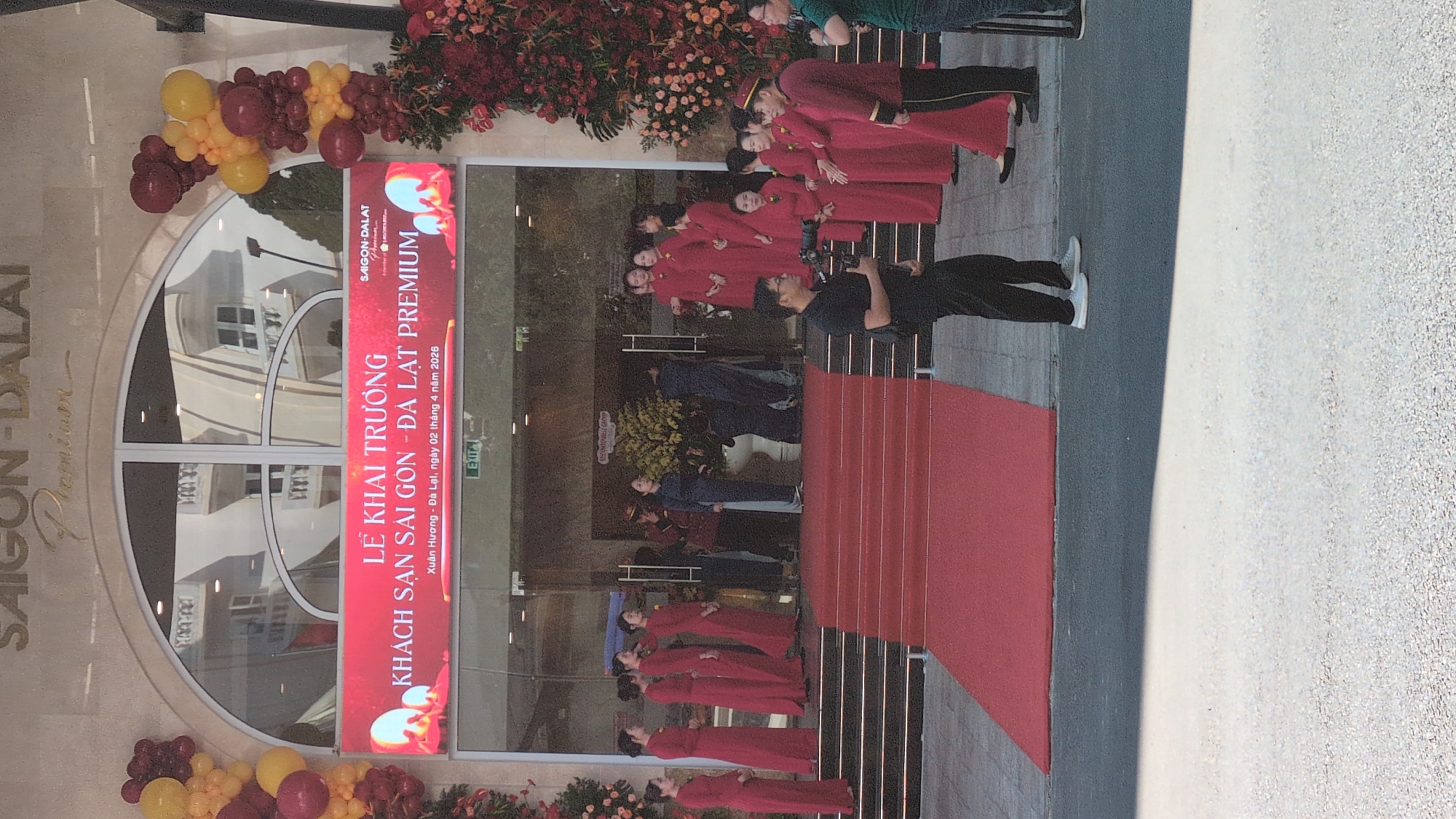 Here is a description of the image:
The image shows the grand opening of the Saigon-Dalat Premium hotel in Da Lat, Vietnam. A large red banner with Vietnamese text announces the opening date as April 2, 2026. A red carpet leads up to the entrance. A group of people in red ao dai (traditional Vietnamese dresses) stand near the entrance. A photographer is positioned nearby, likely documenting the event. Floral arrangements and balloons decorate the entrance. The hotel's name, "Saigon-Dalat Premium," is prominently displayed above the entrance.