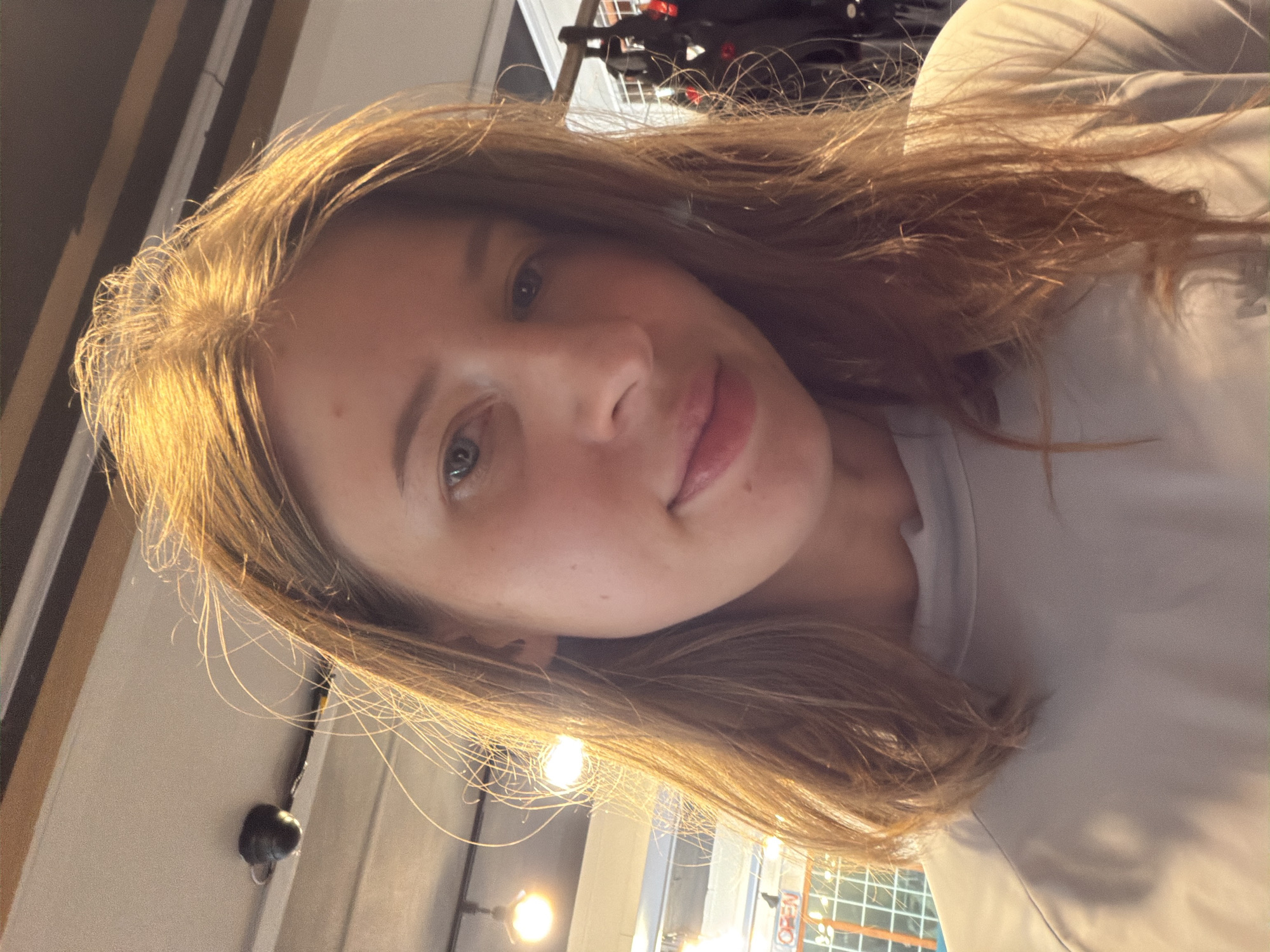 The image shows a young woman with medium-length, light brown hair, taken from a close-up perspective. She is wearing a light grey long-sleeved shirt. The background is blurred but appears to be indoors, with ceiling lights and some racks of dark items visible on the right side. There is no discernible information about the location being in Seo-myeon, South Korea.