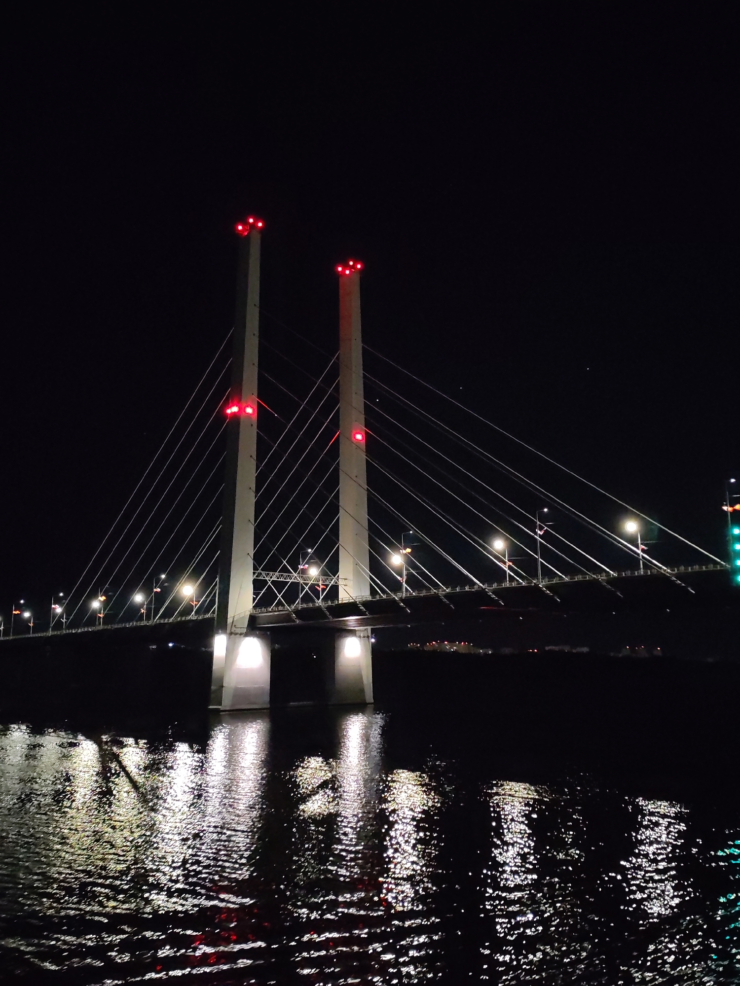 The image depicts the October Bridge in Cherepovets, Russia at night. The bridge's two tall, concrete towers are illuminated with red lights at their peaks and base. Suspension cables extend from the towers to the bridge deck. Streetlights line the bridge, reflecting light onto the water below. The water's surface is dark with bright, scattered reflections of the bridge lights. The background is black, with distant city lights visible along the horizon.