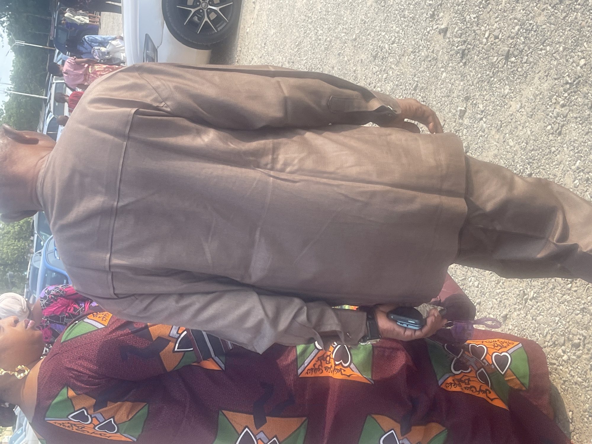 A daytime outdoor scene shows two people in Abuja, Nigeria. The first person, from the back view, is wearing a brown suit. The second person is wearing a maroon and green patterned fabric dress, a white head wrap, and gold earrings. The second person is holding what looks like a smartphone. The setting is a parking area. Additional people, cars, and sparse greenery are visible in the background. The ground is covered with gravel.