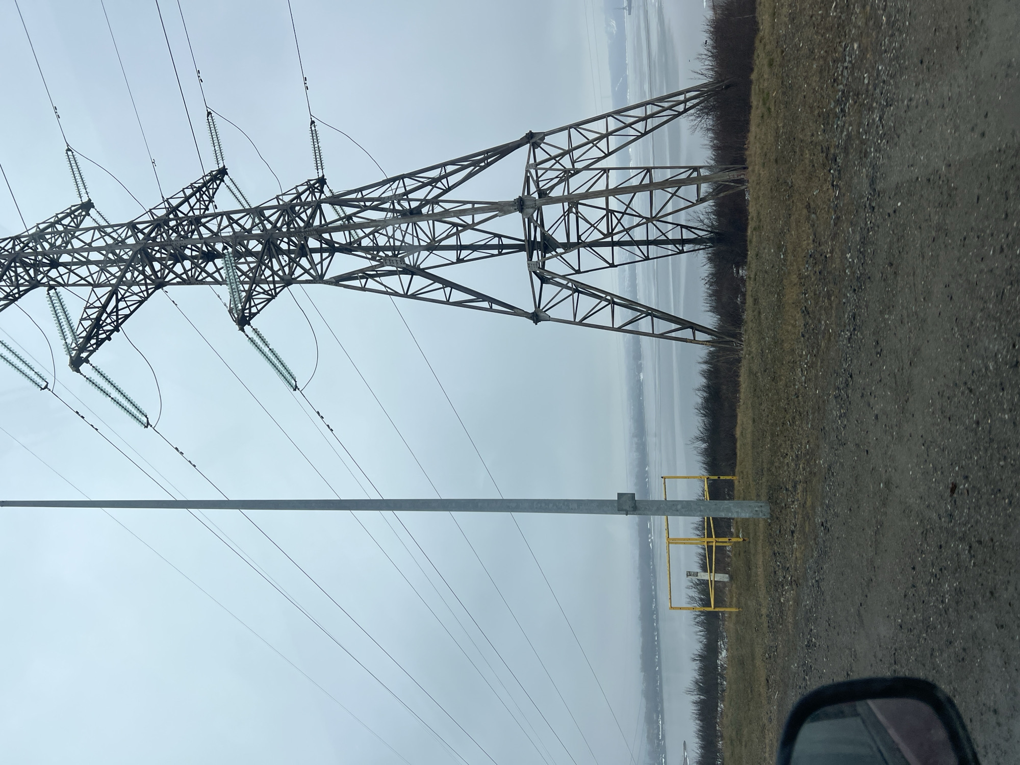 The image depicts a large metal electrical transmission tower alongside a gravel road. Power lines extend from the tower towards the viewer's perspective. A grey metal pole and a yellow guardrail are visible in the mid-ground. A line of trees and a body of water are visible in the background beneath a cloudy sky. The lower-right corner contains the reflection of what appears to be the side-view mirror of a car. The location is Chaikovsky, Russia.