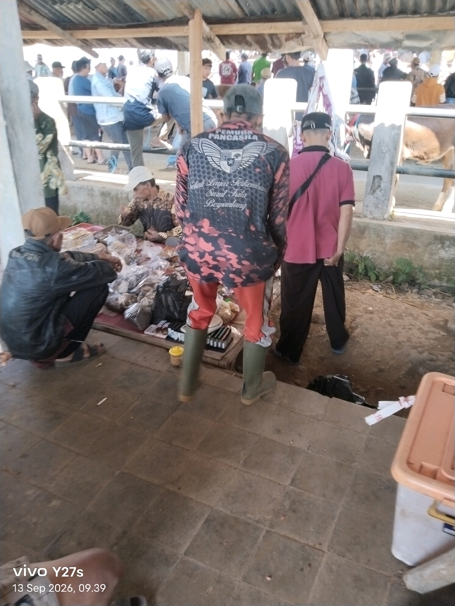 This is a photograph of an outdoor market scene, likely in Wonosido, Indonesia, with a focus on a group of people and stalls. In the foreground, a man wearing a black leather jacket and a brown cap crouches on the ground, next to a stall filled with various food items packaged in plastic. To his right, another man, viewed from the back, wears a dark colored jacket with a red and black camouflage pattern and bright orange pants, along with green rubber boots. He appears to be looking at the stall. Further back, a man in a pink shirt and dark pants stands with his hands in his pockets. In the background, a crowd of people are visible, some standing and some interacting with stalls. A livestock enclosure, possibly for cattle, is visible on the right side of the image. The ground appears to be dirt and concrete. The overall scene depicts a bustling market environment.