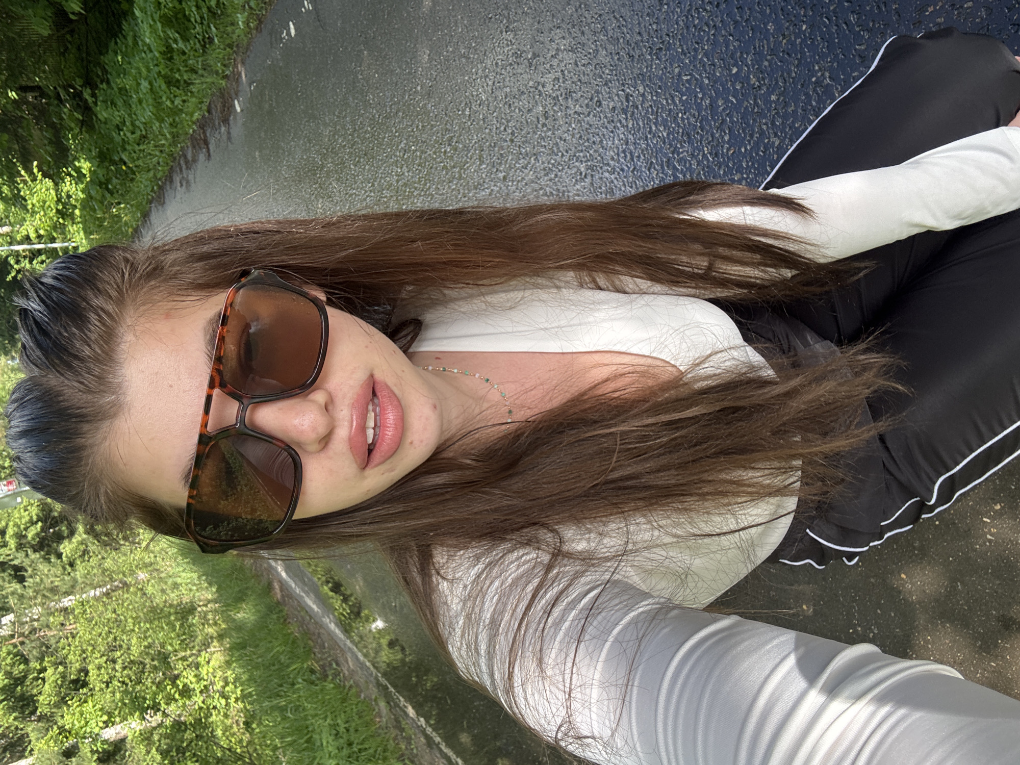 A woman sits on a wet, asphalt surface in Shevlyagino, Russia. She wears sunglasses, a white long-sleeved top, and black pants with white stripes. The asphalt is surrounded by greenery, suggesting a park or roadside location.
