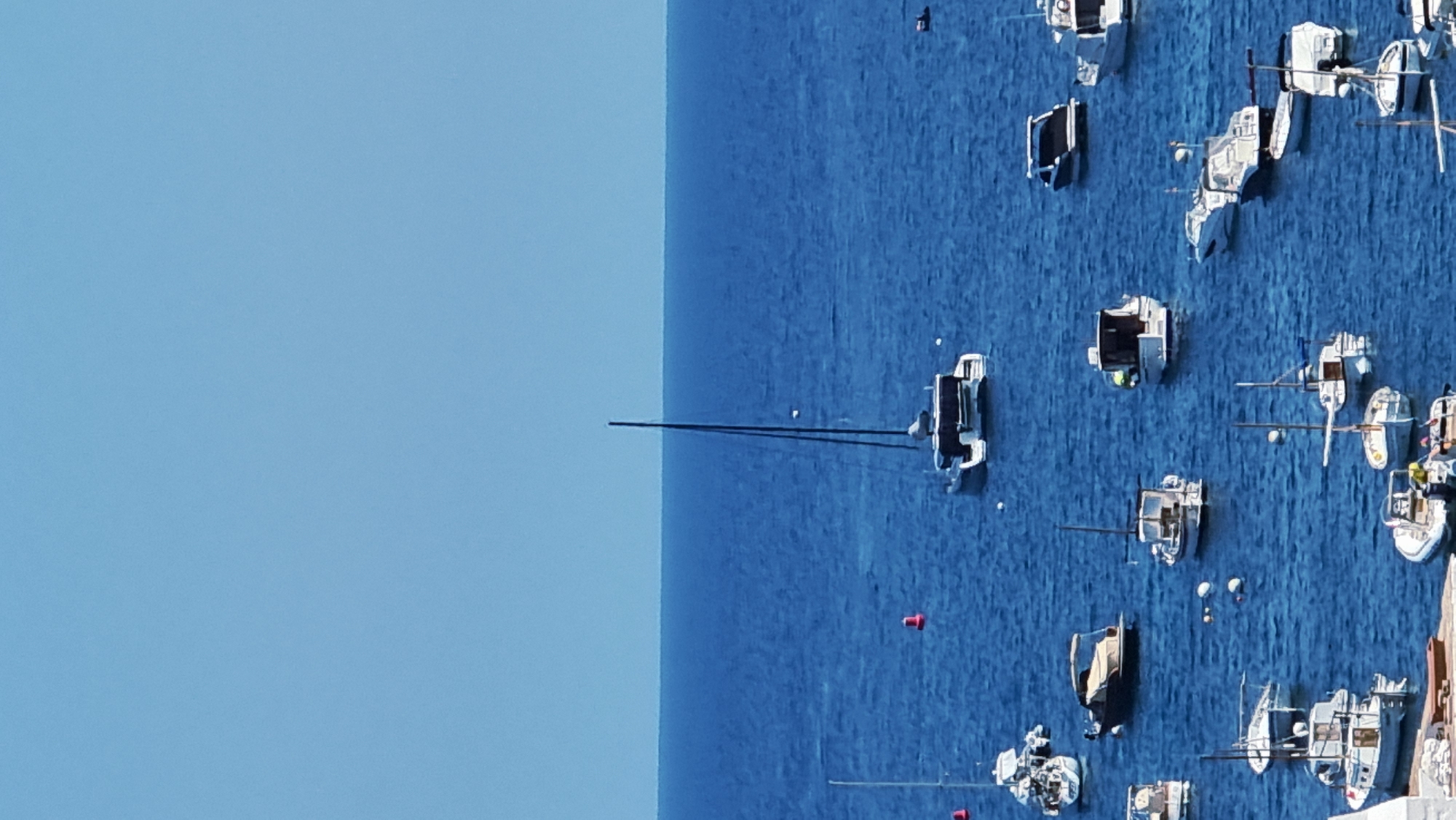 A high-angle, medium shot captures a bay filled with numerous boats of various sizes moored in the deep blue water. In the center of the frame, a large, modern catamaran with two hulls and a tall mast is prominently displayed. Surrounding it are many smaller motorboats and sailboats, some with canopies providing shade. The water appears calm, with gentle ripples reflecting the clear blue sky above. The scene suggests a typical day in a coastal town where recreational boating is popular. The location is the bay of Cadaqués, Spain, known for its picturesque coastline and vibrant maritime activity.