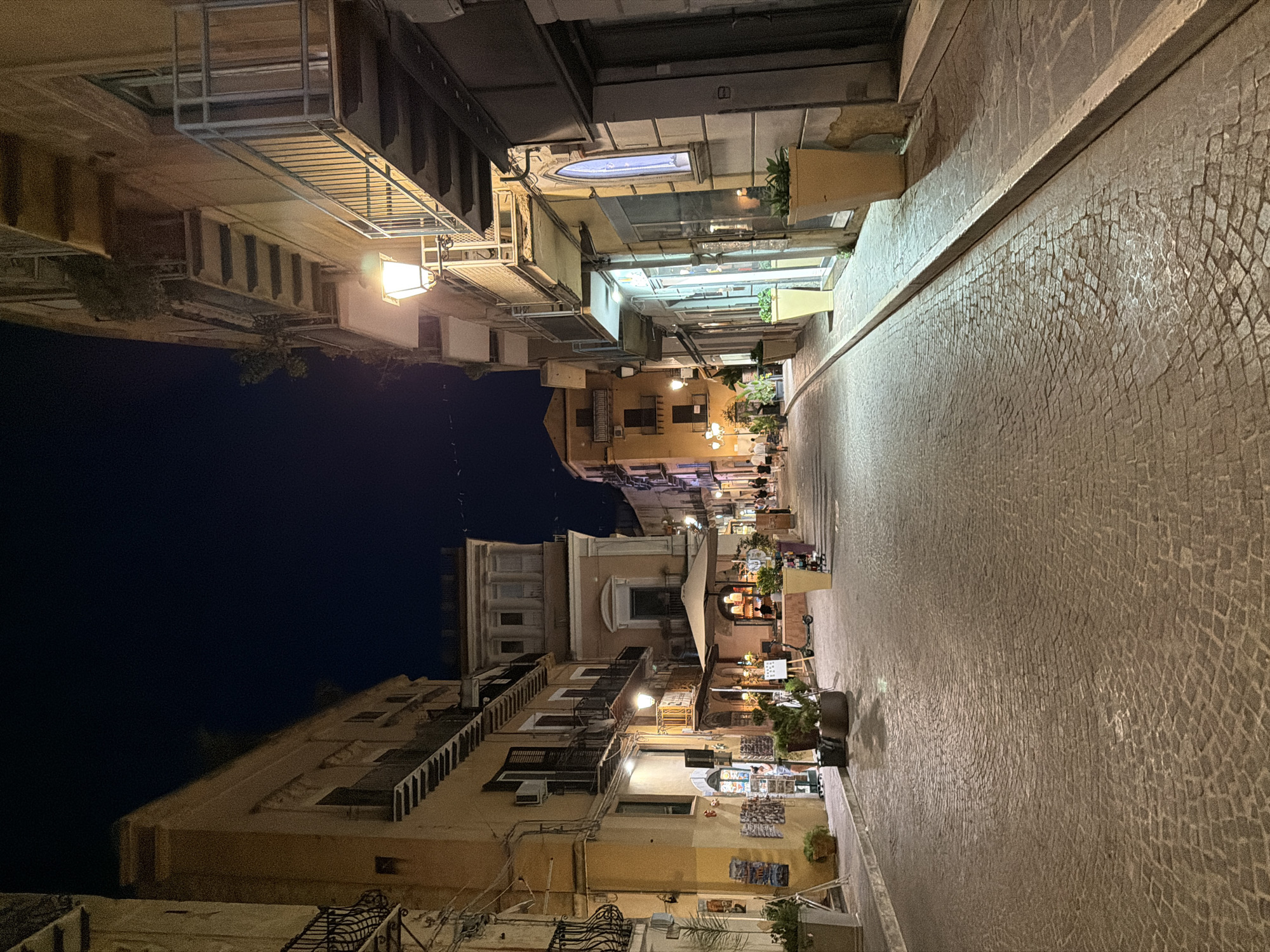 The image depicts a cobblestone street in Agrigento, Italy at night. Buildings line both sides of the narrow street, with balconies and windows visible. The architecture suggests a historic European city. Streetlights illuminate the scene, casting a warm glow on the pavement and building facades. Potted plants are strategically placed along the sidewalks, adding a touch of greenery to the urban landscape. A few people are visible walking in the distance, suggesting a quiet evening atmosphere. The overall impression is one of a charming and atmospheric street scene.