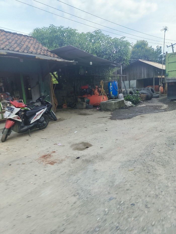 The image shows a dirt road with a small, white motorcycle parked in front of a building with a corrugated metal roof. The building has an open-air structure and a dirt floor. There is a blue barrel with a white label in the background. There is a pile of old tires in the background. The location is a dirt road in the city of Sukatani, Indonesia, with coordinates of latitude -6.617 and longitude 107.406. There are no people in the image.