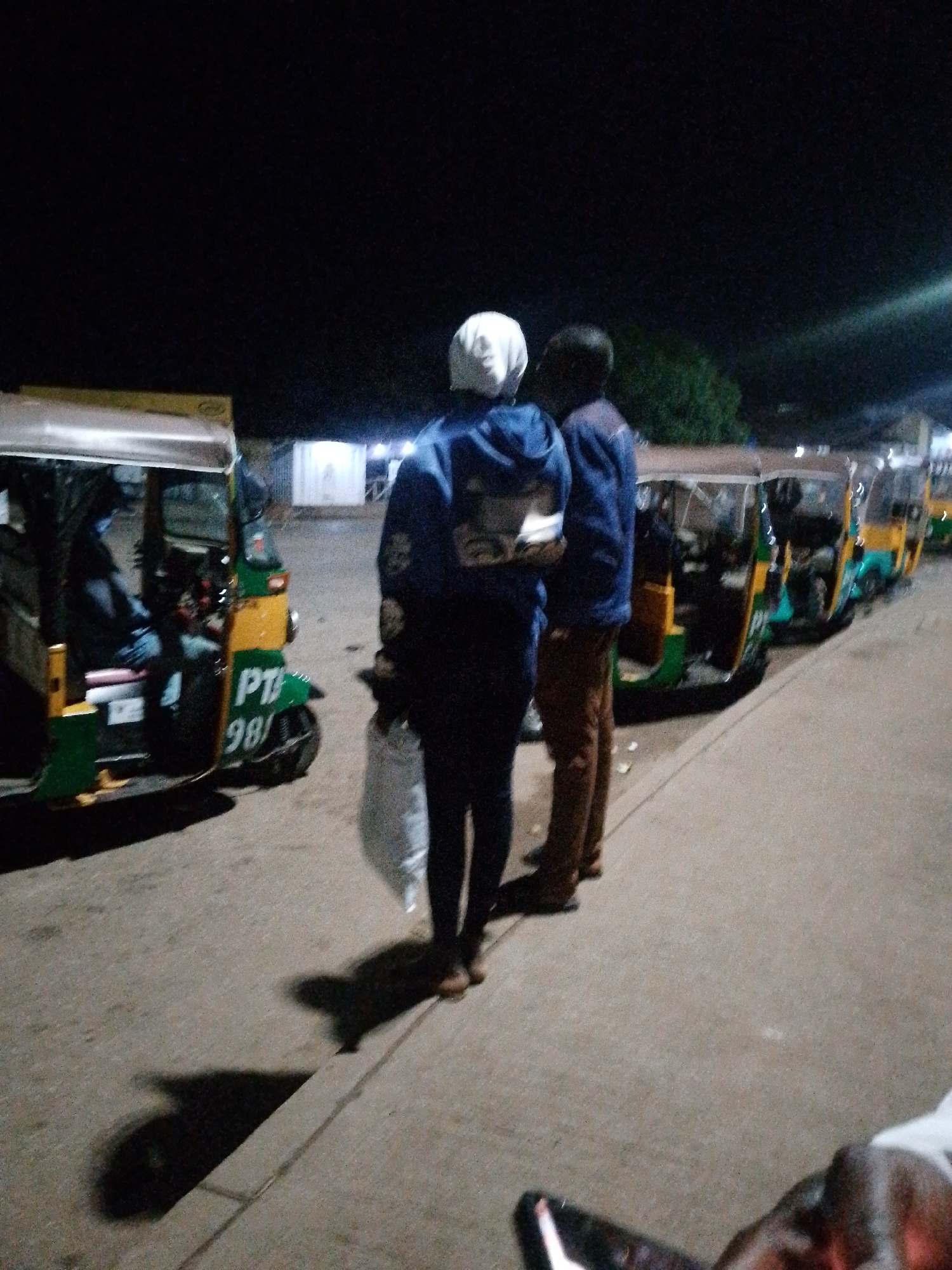 The image shows a group of people standing near a row of auto-rickshaws, also known as "Keke Napeps," at night. Two individuals are prominently featured in the foreground, facing away from the camera. The person on the left wears a white head covering and a blue hooded jacket. The person on the right wears a blue jacket and brown pants. They appear to be waiting or observing the scene.
In the background, several auto-rickshaws are parked in a line. One auto-rickshaw in the foreground has the text "PT 98/" visible on its front. The auto-rickshaws are predominantly green and yellow. The scene appears to be a roadside or a designated waiting area, possibly a motor park, given the number of vehicles.
The lighting suggests it is nighttime, with artificial lights illuminating the area, casting shadows on the ground. The weather is not discernible from the image. The overall atmosphere is calm, with no clear indication of a specific event. The context provided places the scene in Bukuru, Nigeria, suggesting this is a common mode of transportation or a gathering point in that location. The presence of people and vehicles indicates a functional area, likely for transport services.