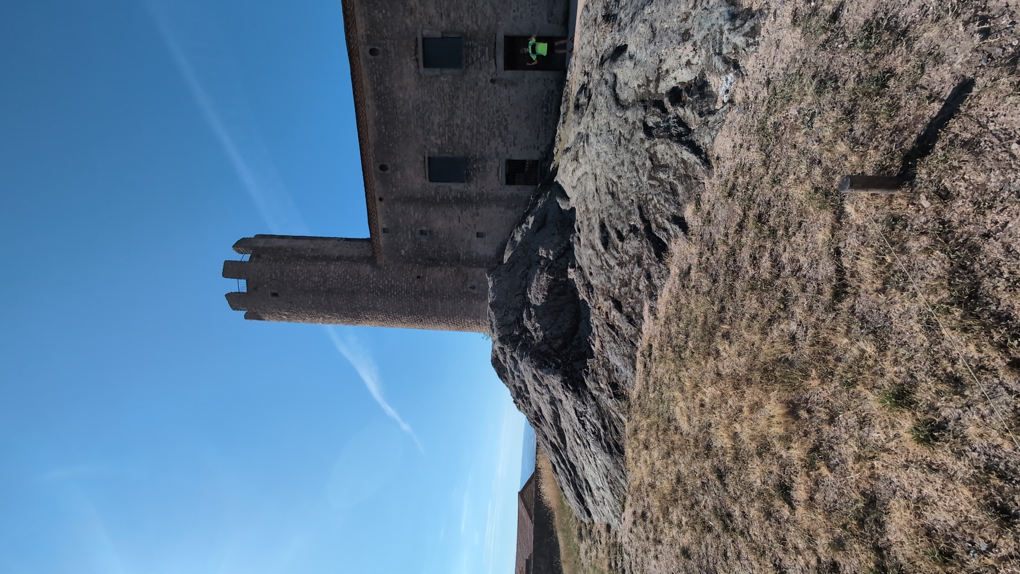 The image depicts the medieval castle of Chambles, France, under a clear blue sky. The castle features a prominent, cylindrical stone tower with crenellated battlements and a long, grey stone building attached to it. The building has small, dark windows and an open doorway where a person in a bright green t-shirt and dark shorts stands, facing away from the camera with their arms raised. The castle is situated on a rocky outcrop, with dry, yellowed grass in the foreground. To the left, another building with a terracotta roof is visible, possibly part of the castle complex or a nearby structure. The overall scene is dry and sunny, suggesting a warm climate.
