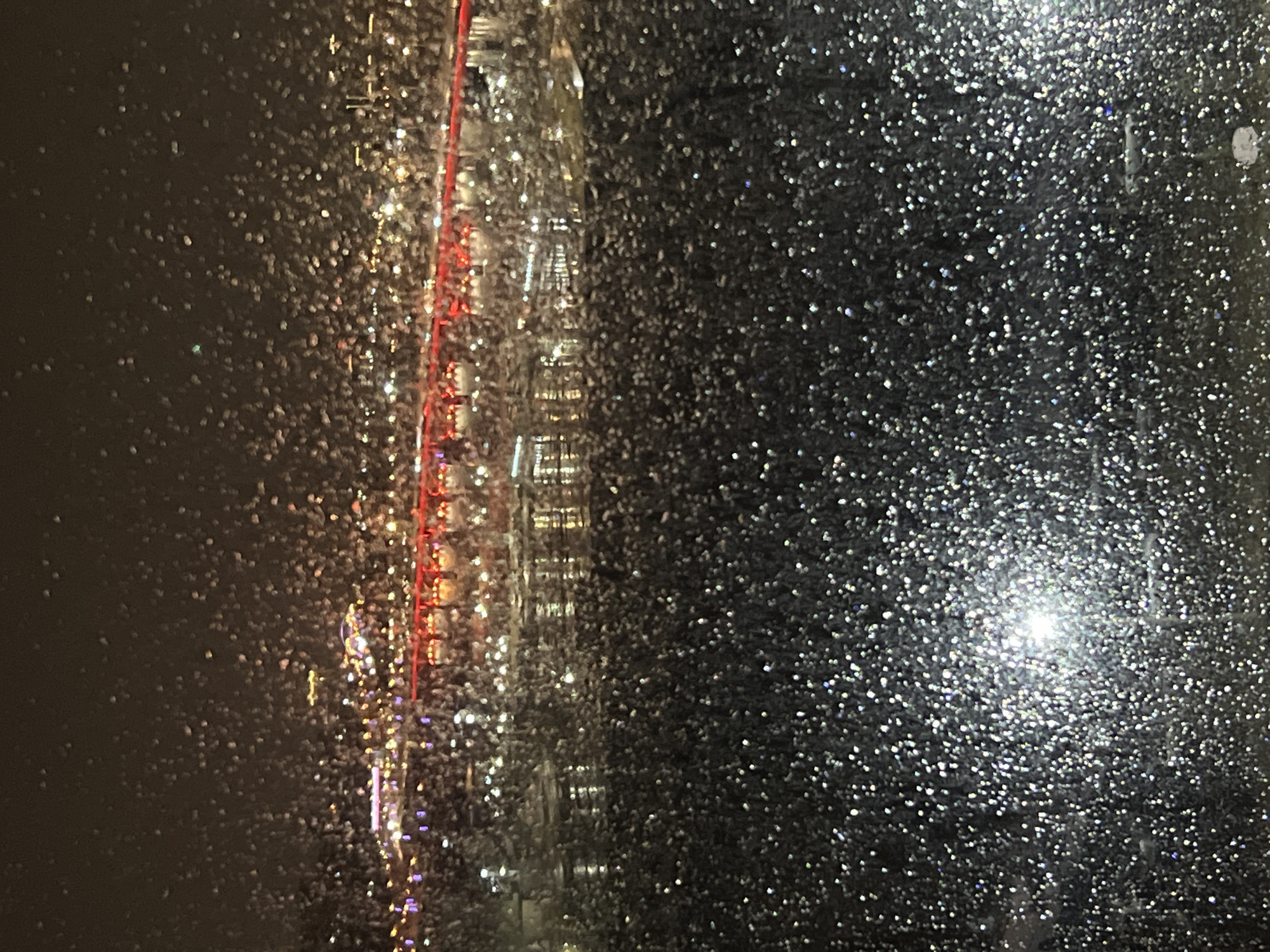 The image appears to be taken through a rain-splattered window at night. On the left, out-of-focus lights and colors suggest urban illumination. A faint, vertical, illuminated structure is discernible in the center. The right portion of the image is dominated by a dense pattern of reflective dots, possibly raindrops on the window, obscuring the background. The bright light in the lower-right suggests a distant light source. The location may be Krasnoyarsk, Russia, but there is no data in the photo to confirm.