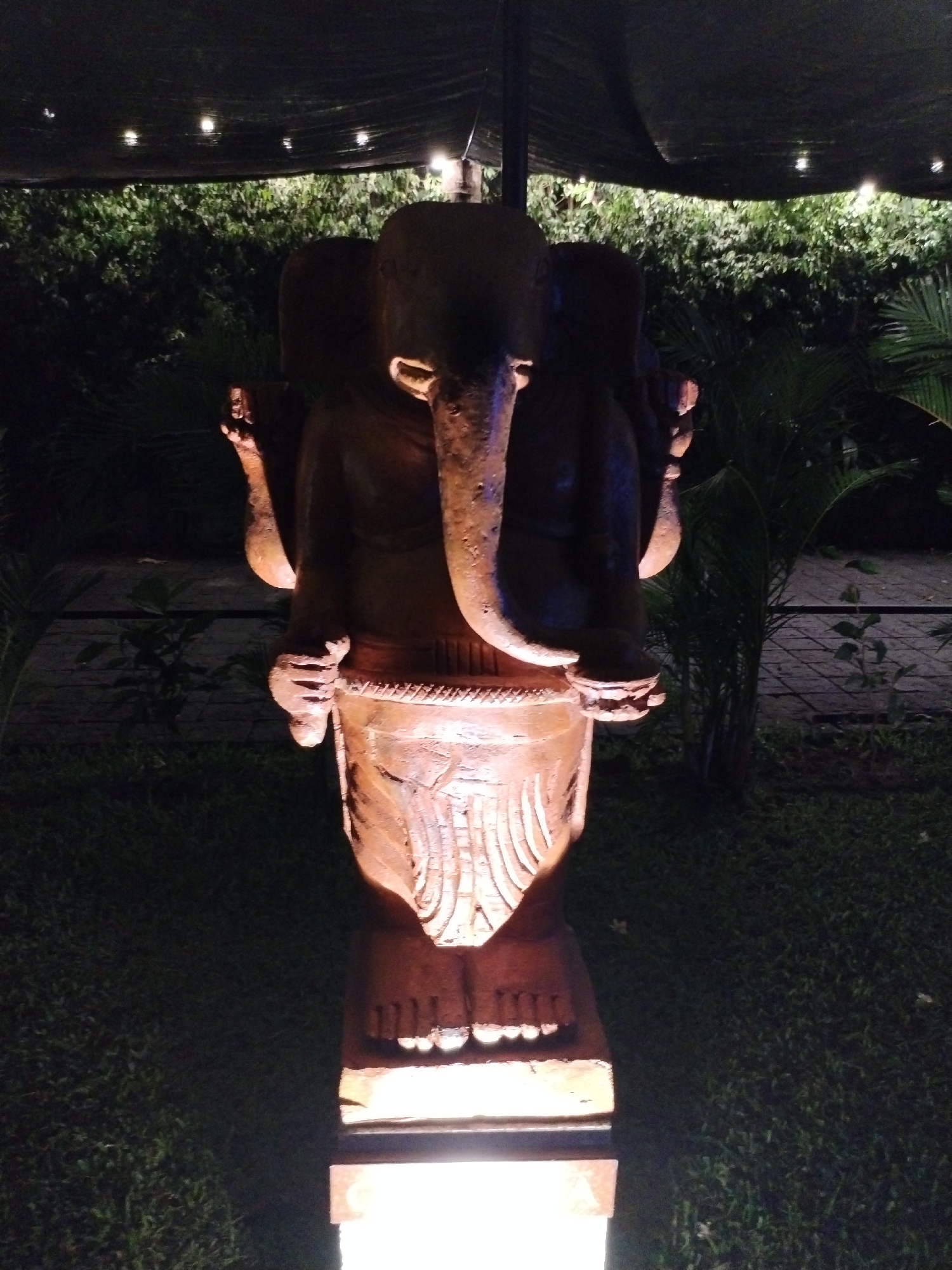 A statue, presumably of Ganesha, is situated on grass in a garden setting, possibly in Bac Nha Trang Ward, Vietnam. The statue has an elephant head with a long trunk and four arms. The bottom part of the statue, below the stomach area, features a set of feet. The statue stands atop a lit pedestal. Above the statue, there is a dark awning with string lights. Behind the statue, there are several green plants, bushes, and trees. The light source for the statue appears to be coming from below.