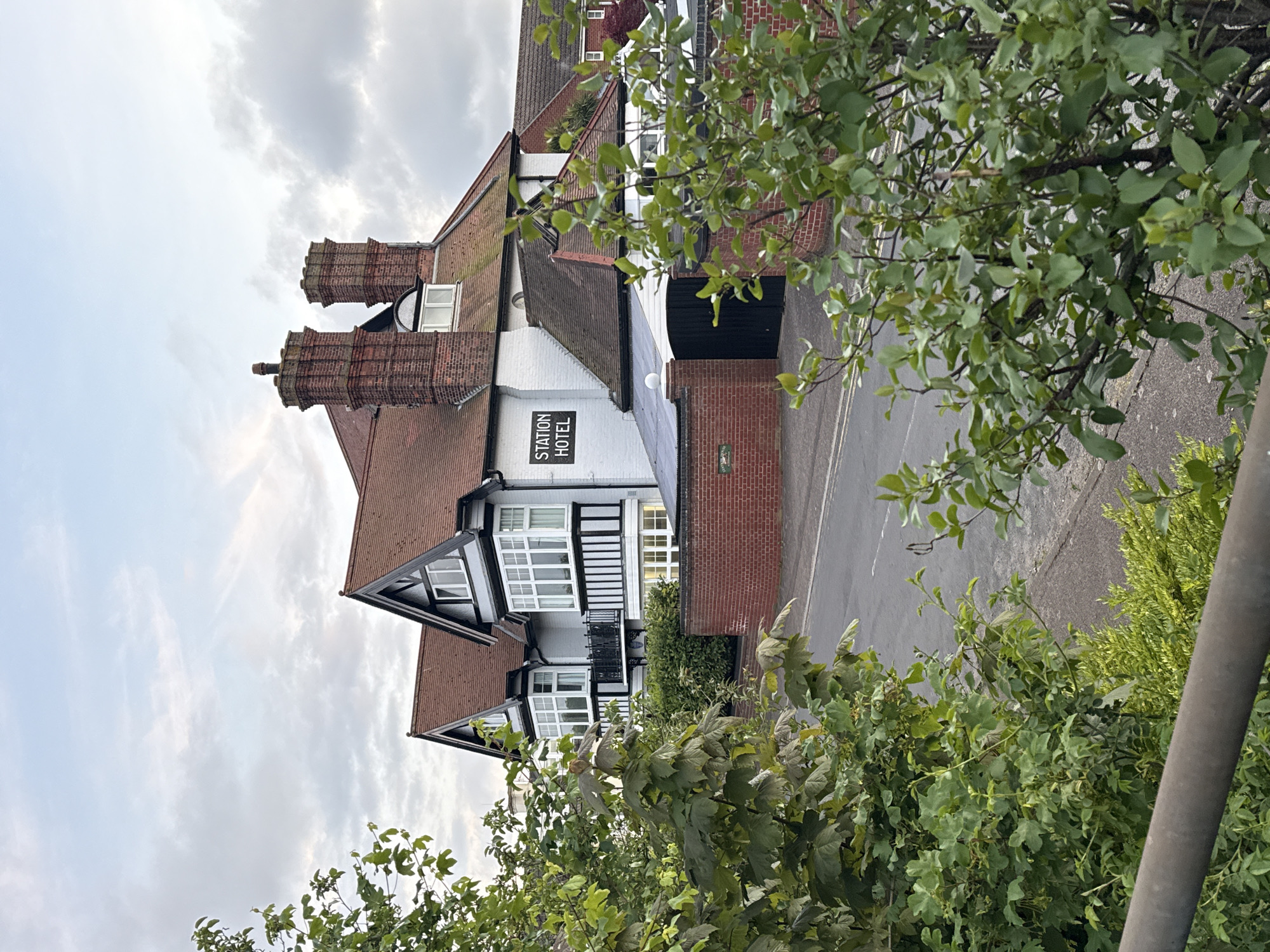 The image depicts the Station Hotel in Great Yarmouth, United Kingdom. The hotel, a multi-story building with a red-tiled roof and white walls adorned with black decorative timbers, is viewed from an elevated position, partially obscured by foliage. Two tall brick chimneys are visible. A black sign reading "STATION HOTEL" is affixed to the building's facade. A paved road runs alongside the building, and the sky is overcast.