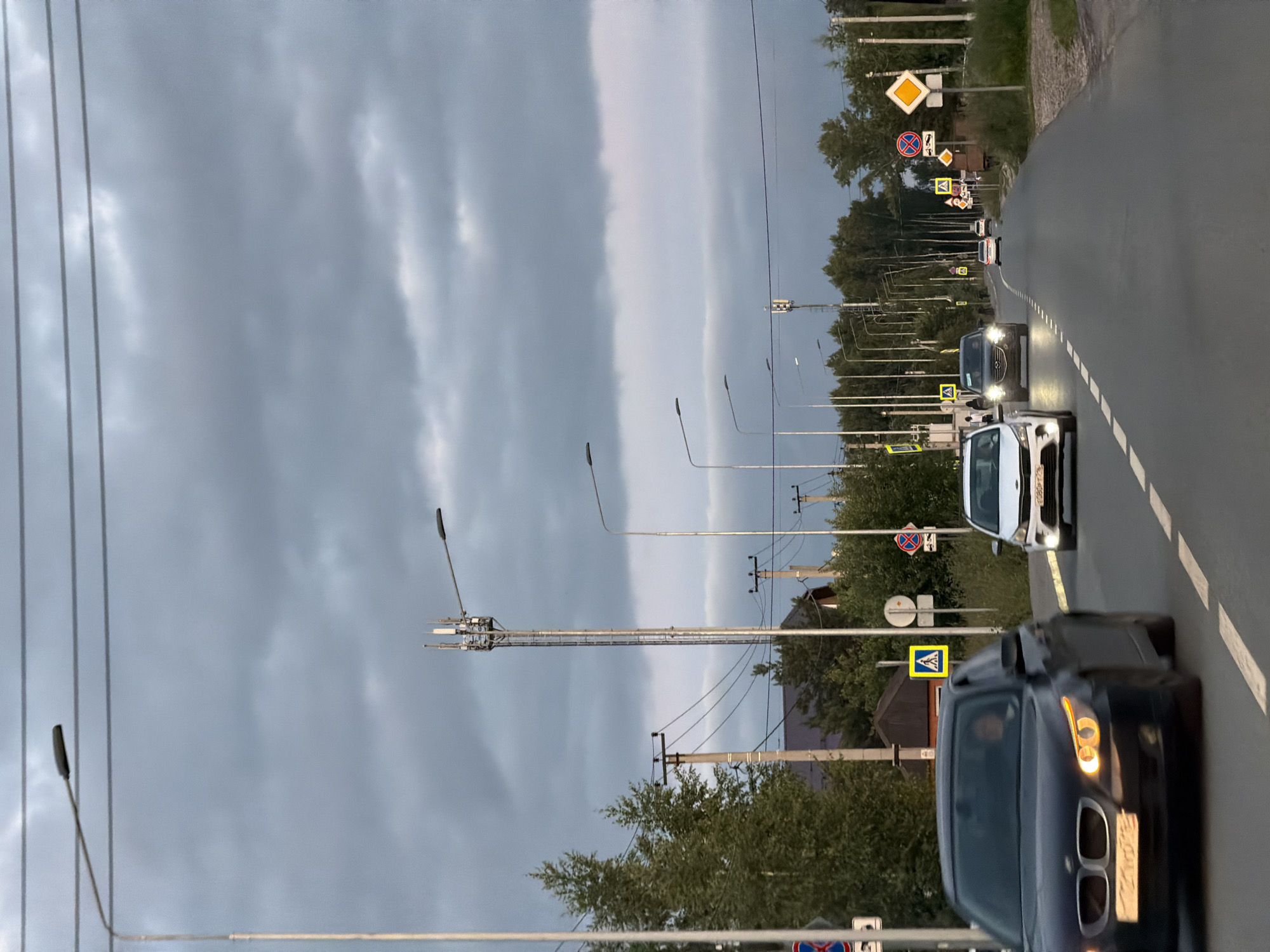 A road with multiple lanes and streetlights runs through a suburban or rural area under a cloudy sky. Several cars are traveling on the road. A dark-colored BMW is in the foreground, with its headlights on. A white car follows behind it, and further down the road, two more cars are visible. On the side of the road, there are trees, power lines, and various traffic signs, including pedestrian crossing signs and a circular sign with a red cross through it. A tall telecommunications tower is also present in the background. The overall scene suggests a late afternoon or early evening setting, possibly in a less densely populated part of Kazan, Russia, given the presence of rural elements like trees and the style of the houses visible.
