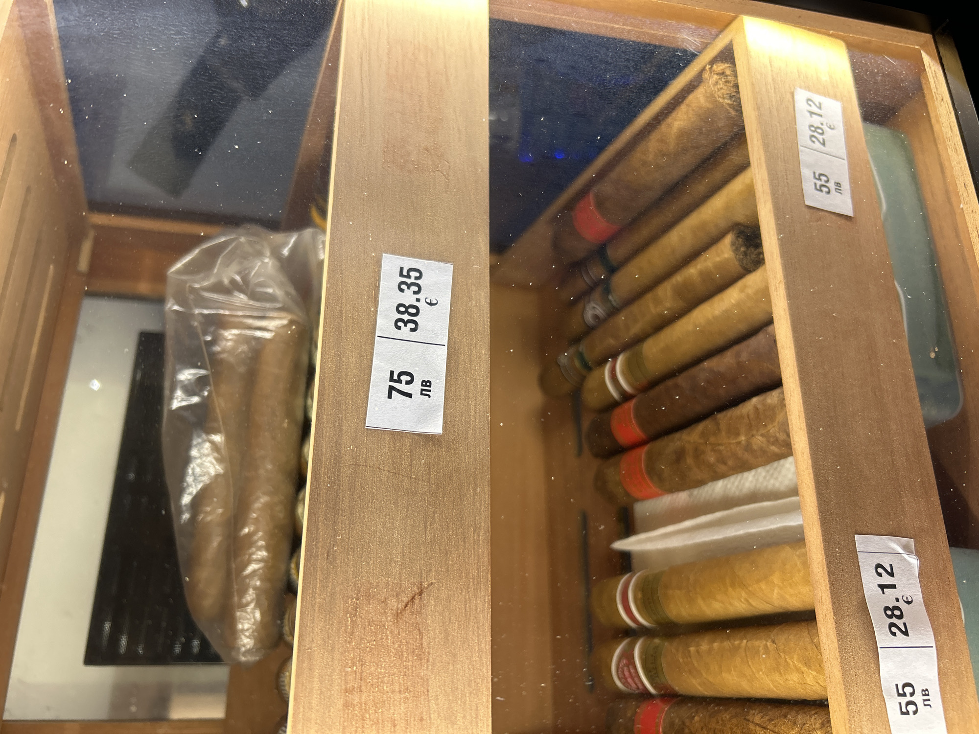 The image shows a close-up of two sections of a humidor, likely located in Plovdiv, Bulgaria, judging by the currency labels on the price tags. The left section contains cigars wrapped in plastic, with a price tag displaying "7.5 лв" and "38.35 €". The right section features a variety of loose cigars, with a price tag showing "55 лв" and "28.12 €". The humidor is made of wood and has a clear lid, allowing a view of the cigars inside. The image appears to be taken in a retail setting, potentially a tobacco shop or a store selling cigars.
