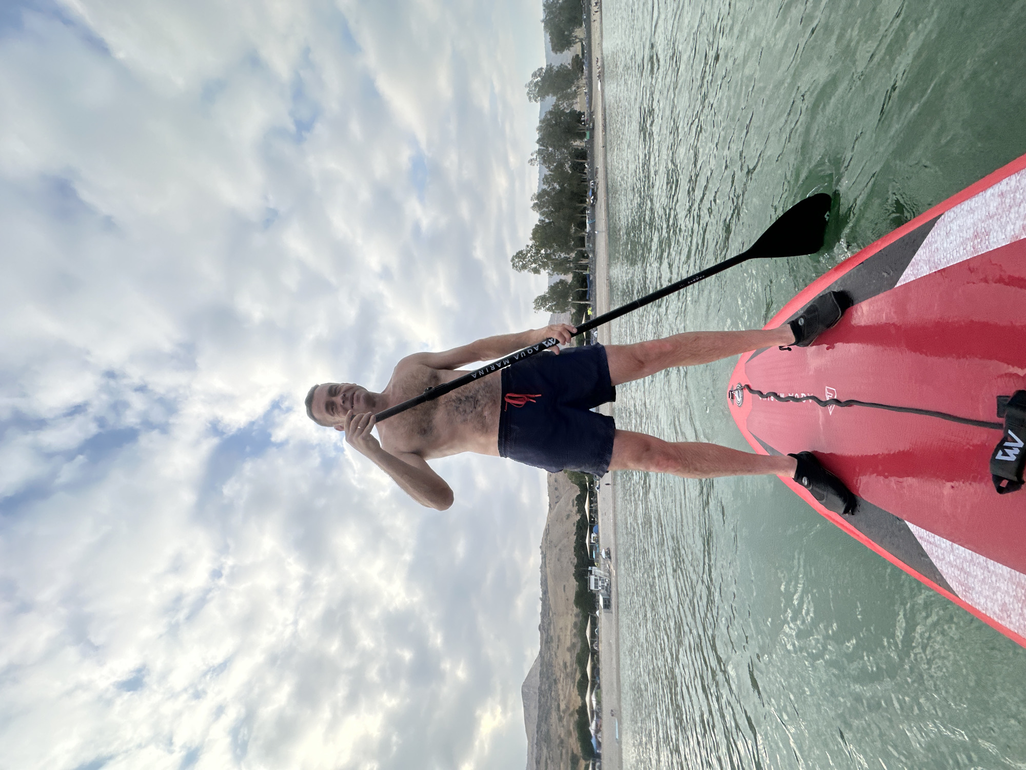 A shirtless man stands on a red paddleboard in a body of water, holding a paddle. He wears dark blue swim shorts and black water shoes. The paddleboard is in the foreground, with its red surface extending towards the viewer. The water is calm with a greenish tint. In the background, a shoreline is visible with a beach, trees, and hills under a cloudy sky. There are a few tents and structures on the beach, suggesting a recreational area. The man is looking towards the camera and smiling. The scene appears to be a scenic location, possibly a lake or sea.