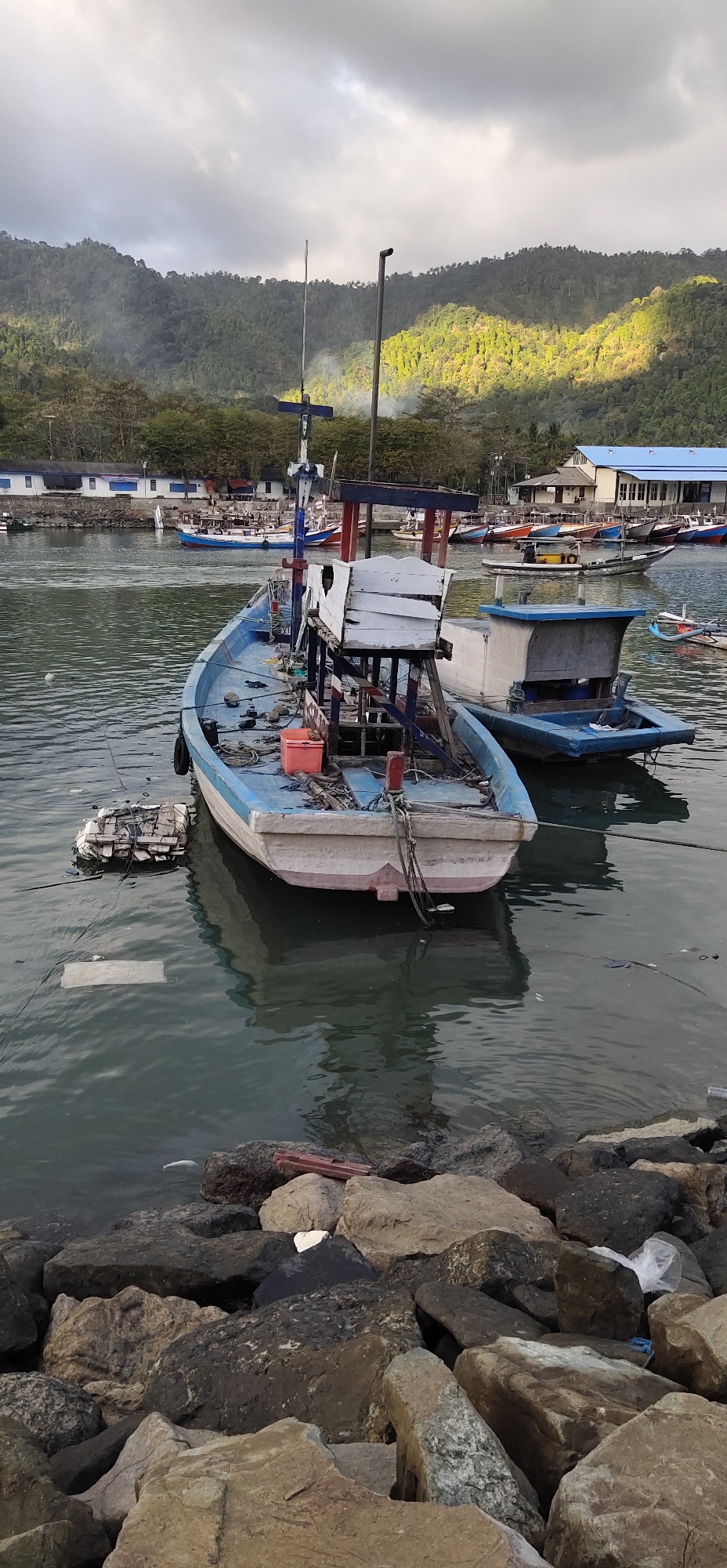 The image depicts a harbor scene in Tasikmadu, Indonesia, with several fishing boats moored in the water. In the foreground, a light blue and white fishing boat is prominently featured, partially submerged with debris floating around it. Another smaller blue boat is tied alongside it. In the background, a line of various other fishing boats are docked, and beyond them, buildings and dense green hills rise under a cloudy sky. The water is calm, with reflections of the boats and the surrounding landscape visible. The rocky shoreline in the immediate foreground is littered with some trash, including plastic bags and debris. The lighting suggests it is either early morning or late afternoon, with sunlight filtering through the clouds and illuminating parts of the distant hills.