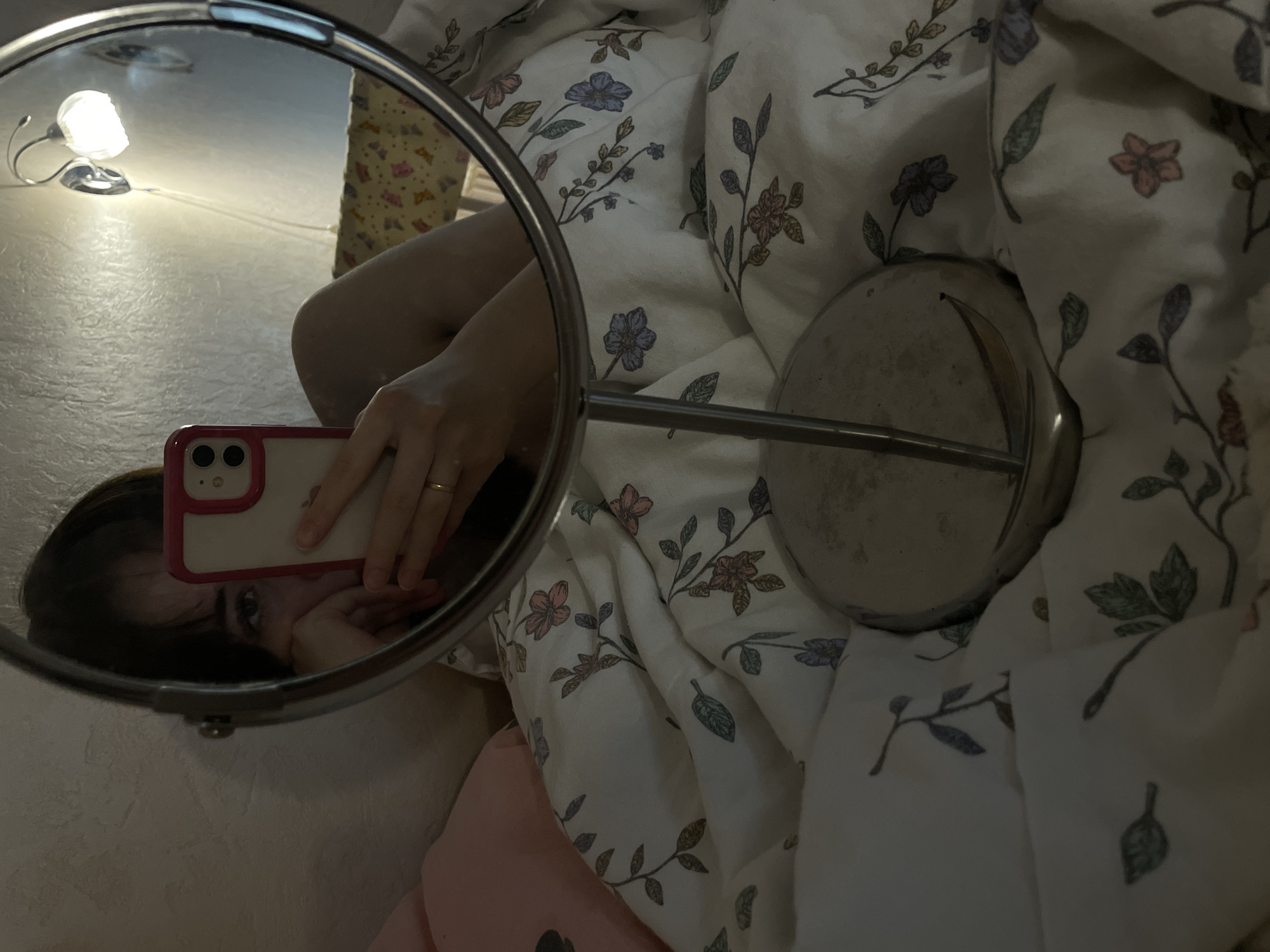 A person is taking a selfie in a round mirror. The mirror reflects the person's face, a white smartphone with a pink case, and a desk lamp. The mirror's base and stem are visible. The background consists of a white floral-patterned comforter. The image suggests an indoor location, likely a bedroom. There's no information available to confirm the city or location of Chaikovsky, Russia.