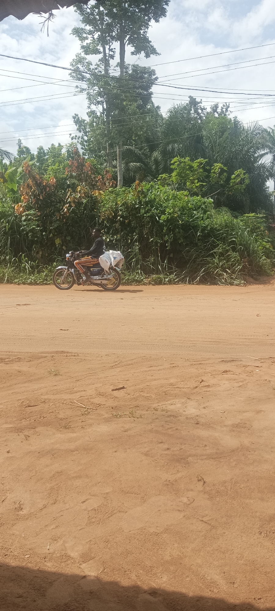 A man is riding a motorcycle on a dirt road in Amauzu, Nigeria. The man is carrying packages on the motorcycle. The background consists of trees, vegetation, and power lines against a cloudy sky. The ground is brown dirt.