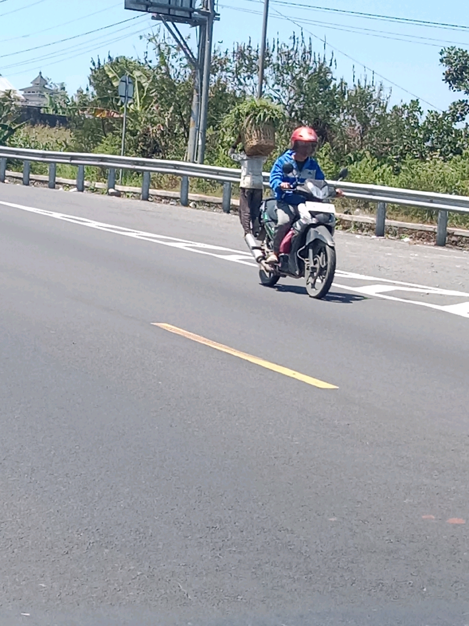 A man wearing a red helmet and a blue jacket rides a scooter on a paved road in Purwojati, Indonesia. He is on the right side of the road. Behind him, another person is walking, carrying a basket of plants on their head. The road is bordered by a metal guardrail and various trees and vegetation. A building with a distinct architectural style, possibly a temple or mosque, is visible in the background on the left side of the image. The sky is clear and blue.