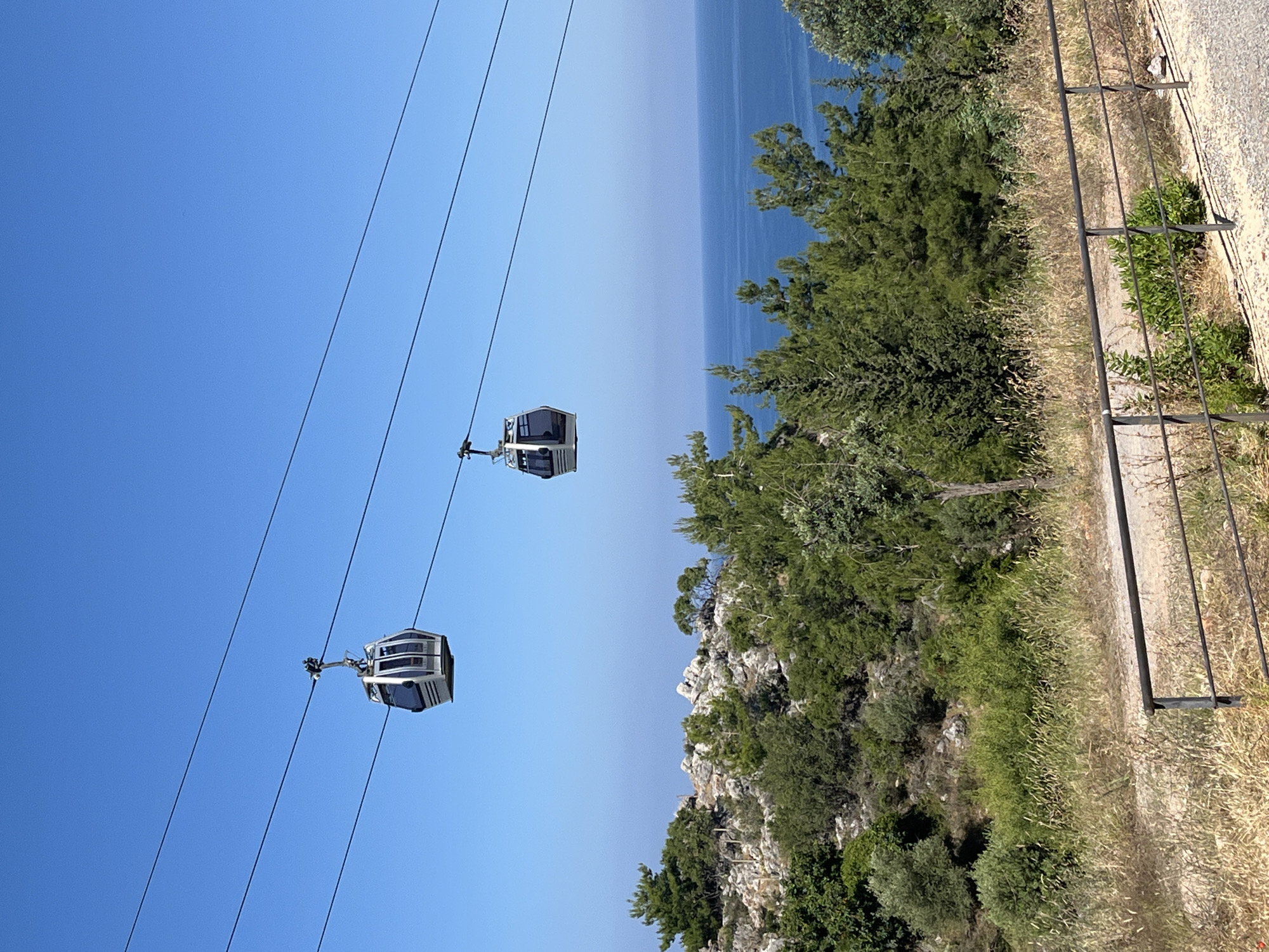The image shows a mountainside view in Alanya, Turkey. Two cable cars are suspended on cables against a clear blue sky, moving along the line. The Mediterranean Sea is visible in the background, partially obscured by trees on the mountainside. The mountainside is covered with dense green trees and shrubs. A metal fence is situated on the edge of a path. The ground near the path consists of dry grass and dirt.