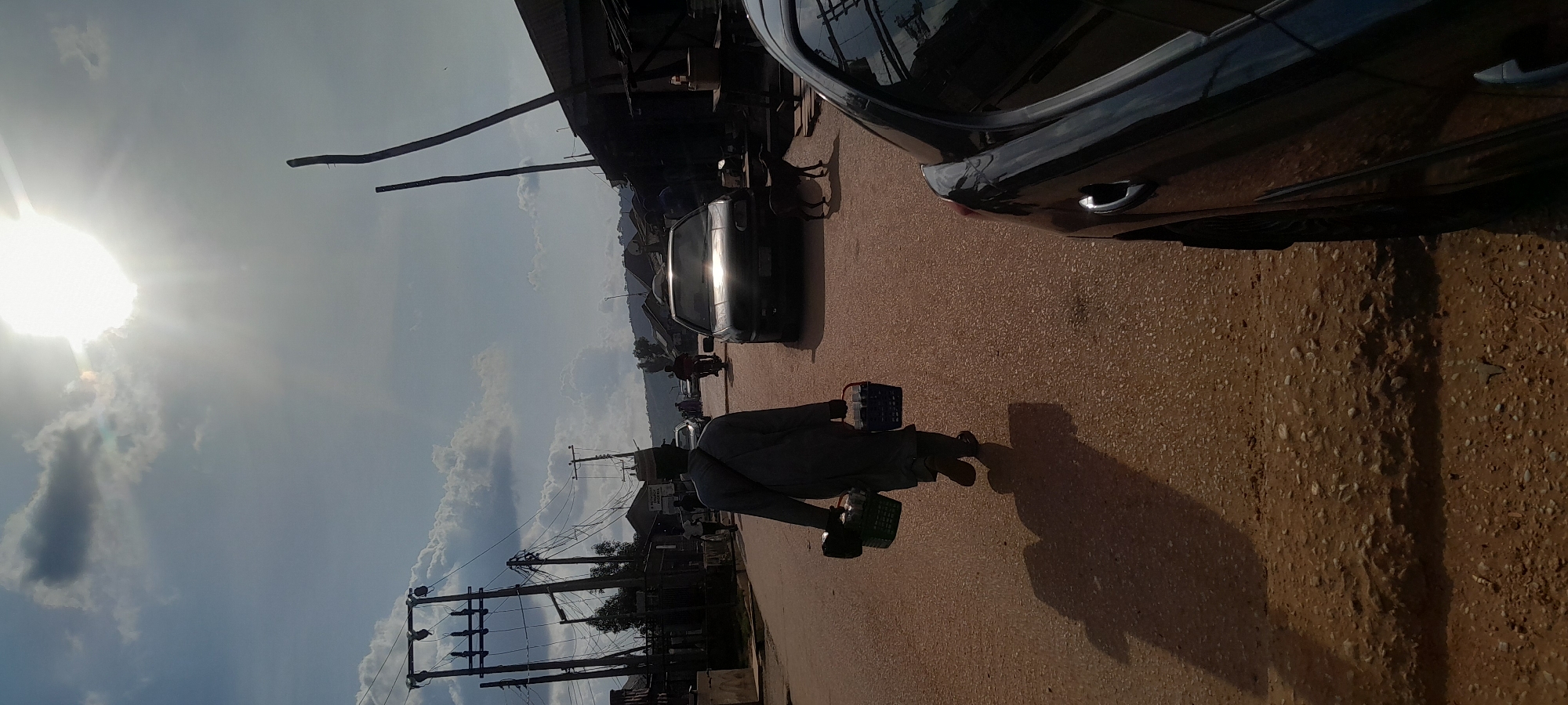 A full shot depicts a person walking away from the camera on a dirt road, carrying two baskets. The person is wearing a gray garment and a hat, and their shadow is cast on the road. The scene is set in Abuja, Nigeria, under a bright, sunny sky with scattered clouds. Several vehicles, including a car and a motorcycle, are visible on the road, along with power lines and buildings on the sides of the street. A dog stands near the side of the road. The overall impression is of a typical day in a Nigerian town.