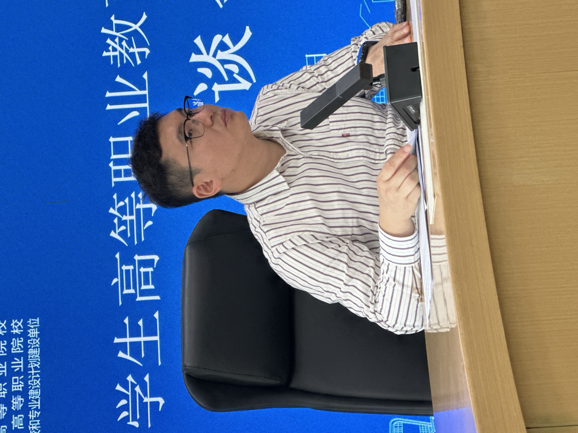 The image depicts a man seated at a wooden desk in an indoor setting, likely in Jiangjin, China. He is wearing glasses and a light-colored striped collared shirt. He looks attentively upwards and to his left (from his perspective), with a neutral expression. His right hand rests near a black, microphone-like device on the desk, while his left hand is on some papers. He is seated in a black office chair.
Behind the man is a large blue backdrop or screen featuring white Chinese characters. The visible text, when oriented correctly, includes:
- "高等职业院校" (Higher vocational colleges)
- "和专业建设计划建设单位" (and professional construction planning and construction unit)
- "学生高等职业教" (Students' Higher Vocational Education)
- "谈。" (talk/discussion)
The setting suggests a conference, lecture, or educational event related to vocational training or academic discussions. The man appears to be a participant, listener, or perhaps a speaker at this event. The lighting is bright, indicative of an indoor daytime scene.