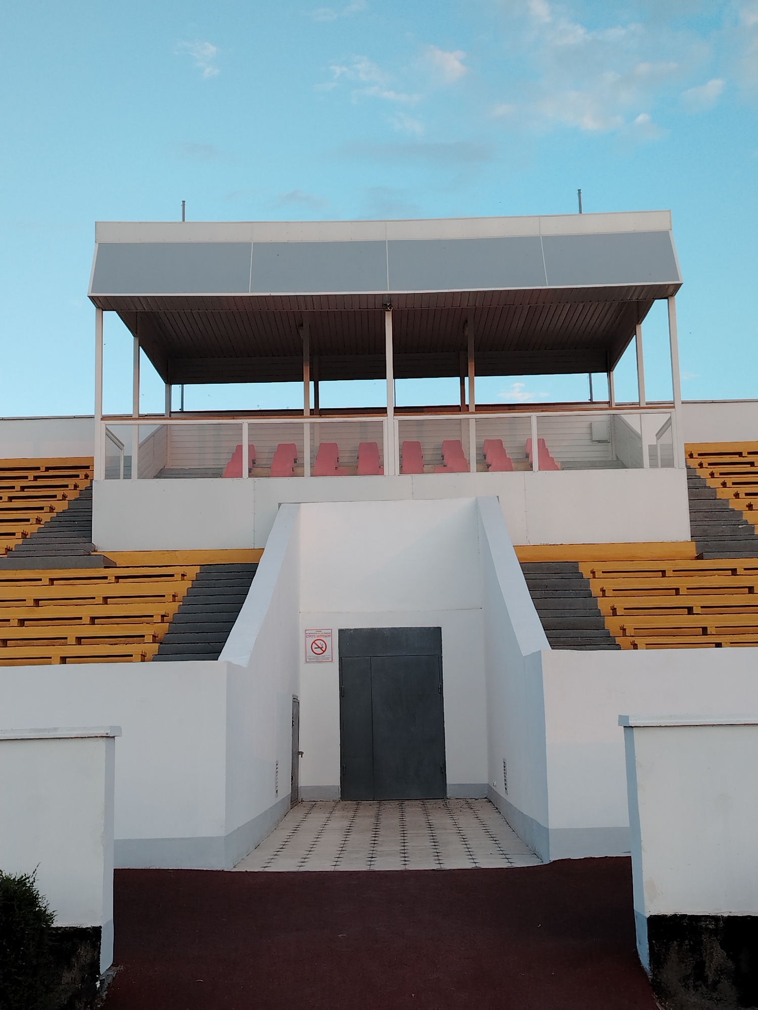 This is an outdoor, eye-level, medium shot of a stadium grandstand with a blue sky and scattered clouds above. The grandstand is predominantly white with yellow seating and grey steps. A covered section with a grey roof and wooden ceiling sits at the top, with a row of red seats visible behind a glass partition. Below this, the white structure of the grandstand features a central entrance with a grey door. A sign with a red circle and slash over a cigarette is visible to the left of the door. The area in front of the entrance is paved with checkered tiles. The stadium is situated in Baikonur, Kazakhstan. The lighting suggests it is late afternoon or early evening.
