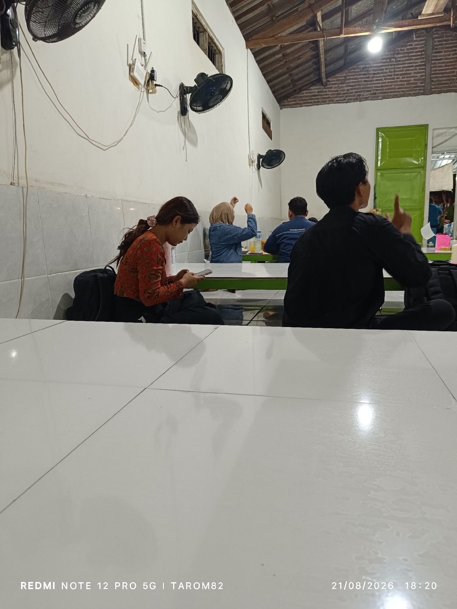 In Sidodadi, Indonesia, an indoor scene depicts a group of people seated on the floor at tables. A woman in a red patterned top and black pants is in the foreground, looking down at her phone. Behind her, a woman in a denim jacket and headscarf is seated, with her hands raised. A man in a black shirt is in the mid-ground, with his back to the camera, gesturing with his right hand. Another person in a blue shirt is seated further back. The environment appears to be a dining area with white walls, a wooden ceiling, and a green door. Two ceiling fans are visible, one on the left and one on the right. The floor is tiled in white. A timestamp at the bottom reads "21/08/2026 18:20."