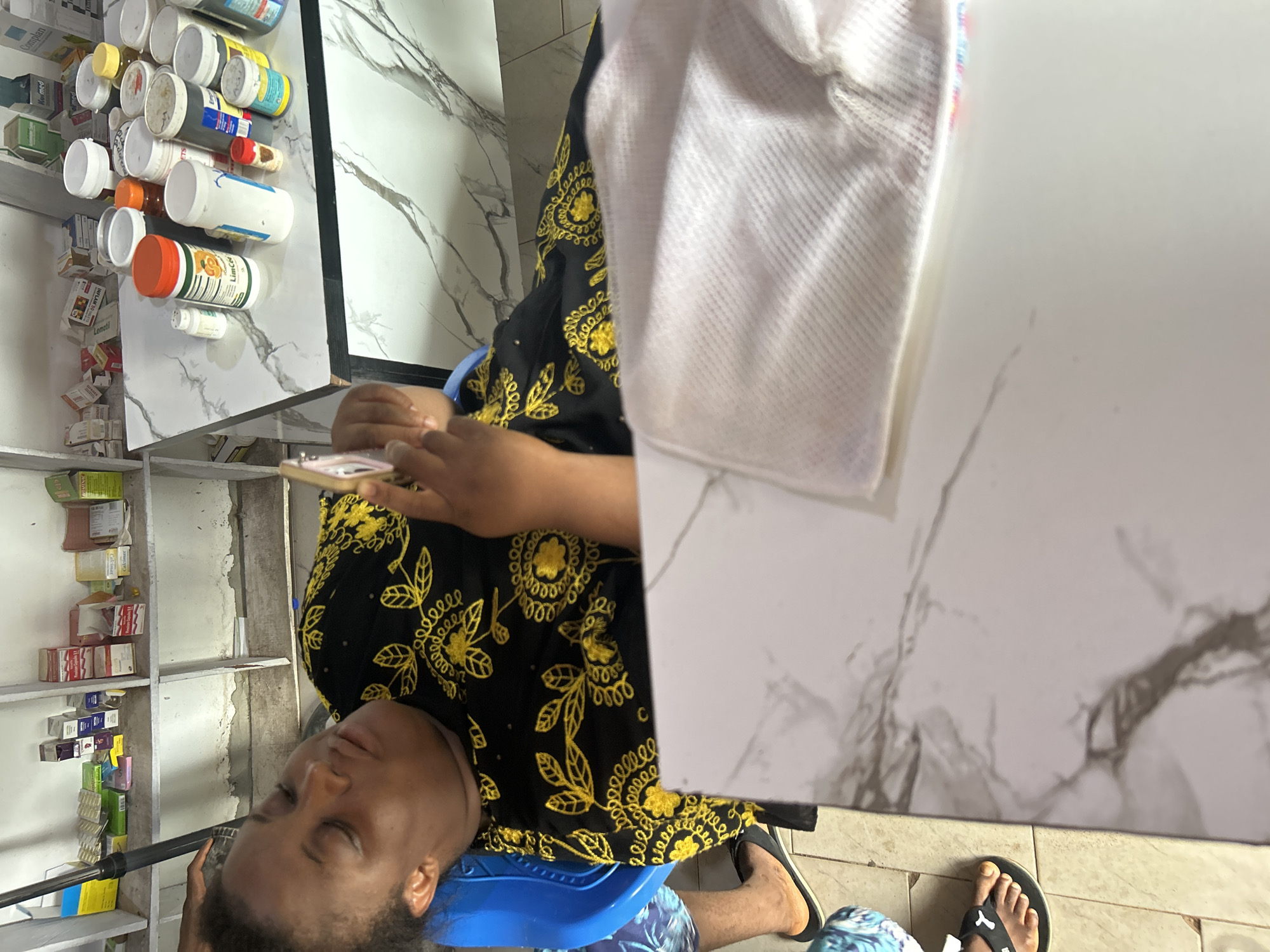 A woman sits on a blue plastic chair in Ihiagwa, Nigeria, holding a smartphone. She is wearing a black dress with gold embroidery. She is barefoot, wearing black flip-flops. The location appears to be a small pharmacy or shop, with shelves stocked with medications and various products. Various pill bottles are visible on the counter above her.